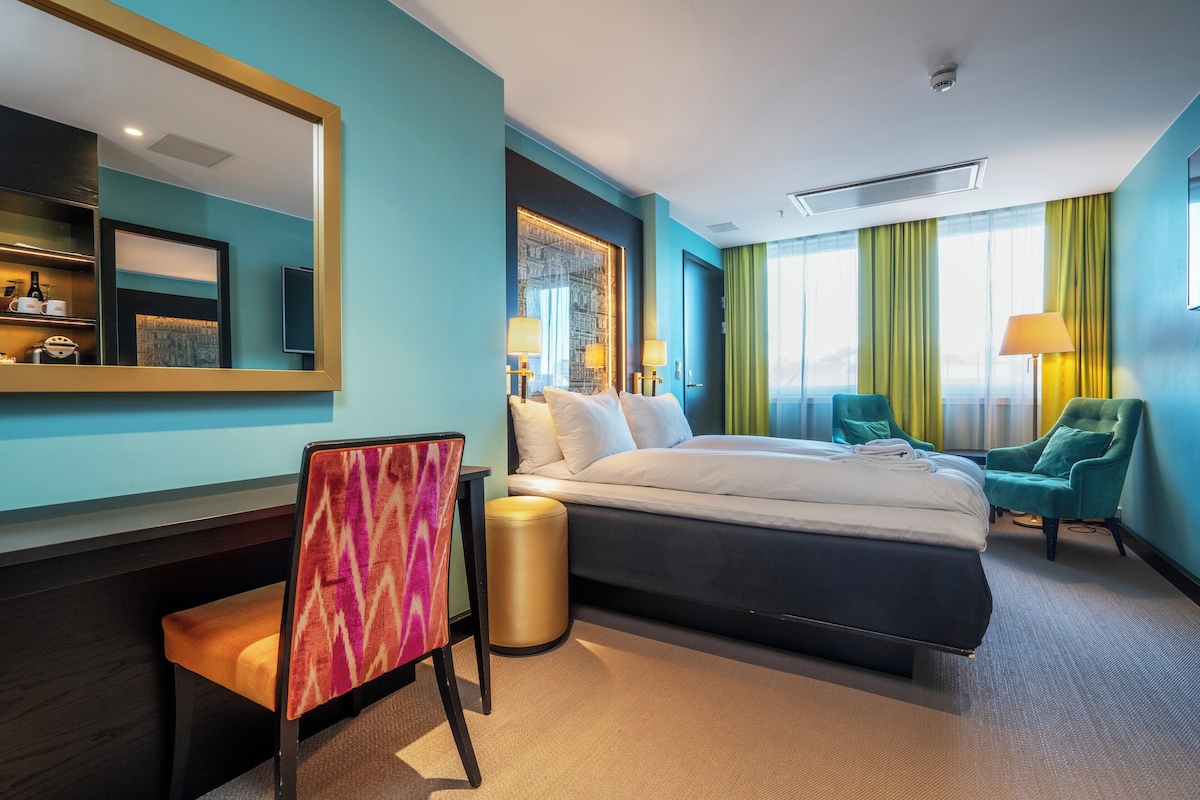A hotel room with a bed, a desk, a chair, a trash bin, and a large mirror.