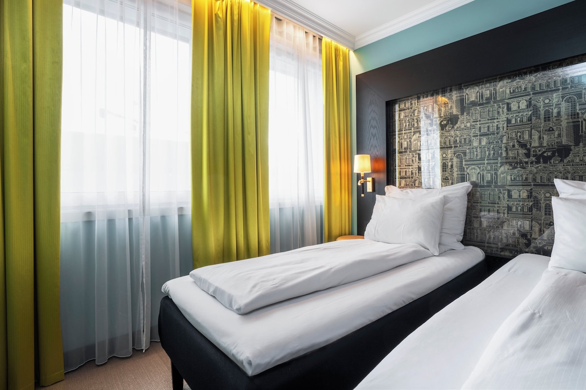 A hotel room with a bed, curtains, a lamp, and a wall with a detailed mural.