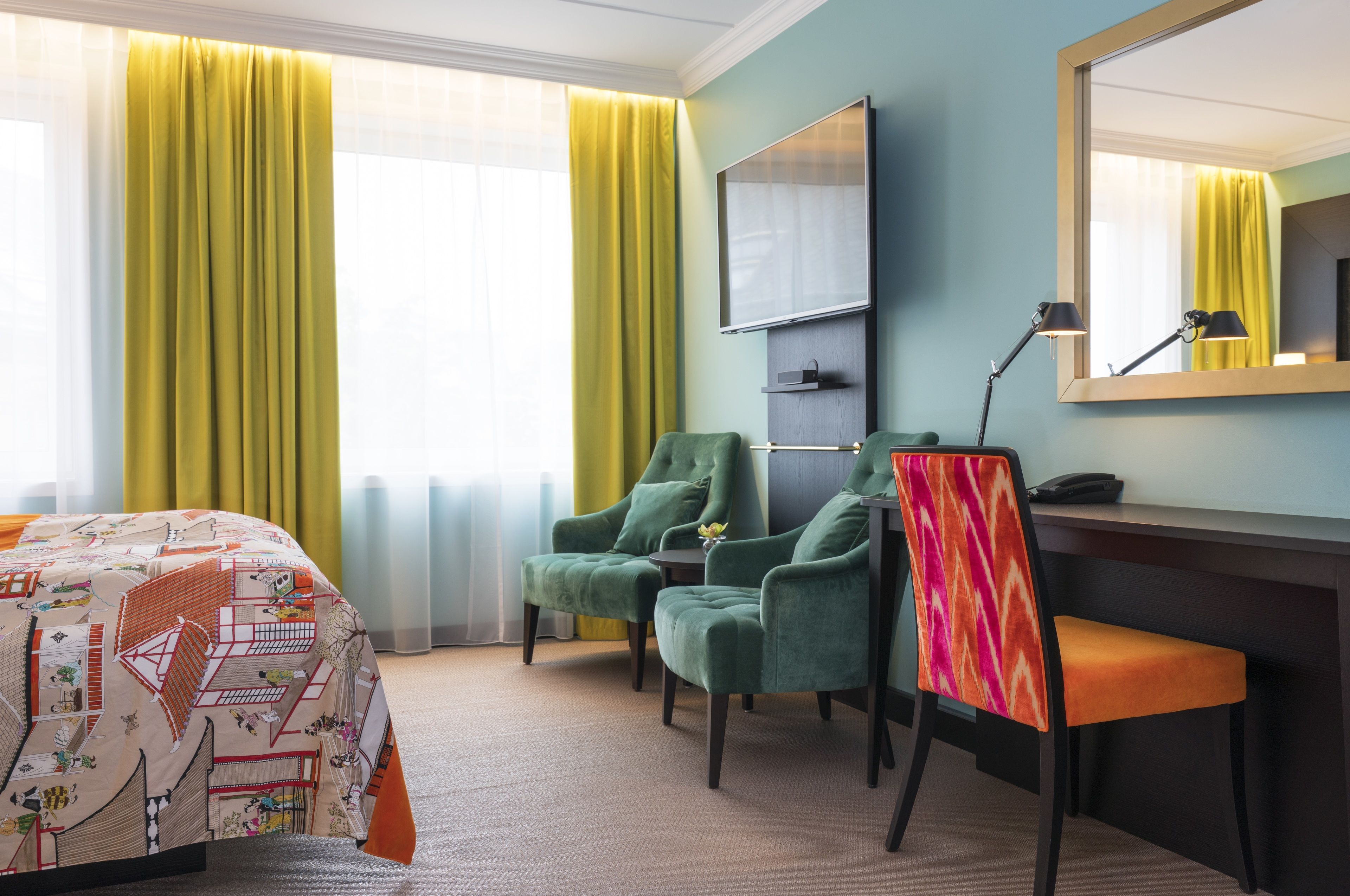 Hotellihuone, jossa on sänky, kaksi tuolia, työpöytä, peili ja ikkuna, jossa on verhot.