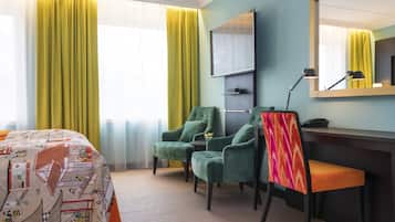 A hotel room with a bed, two chairs, a desk, a mirror, and a window with curtains.