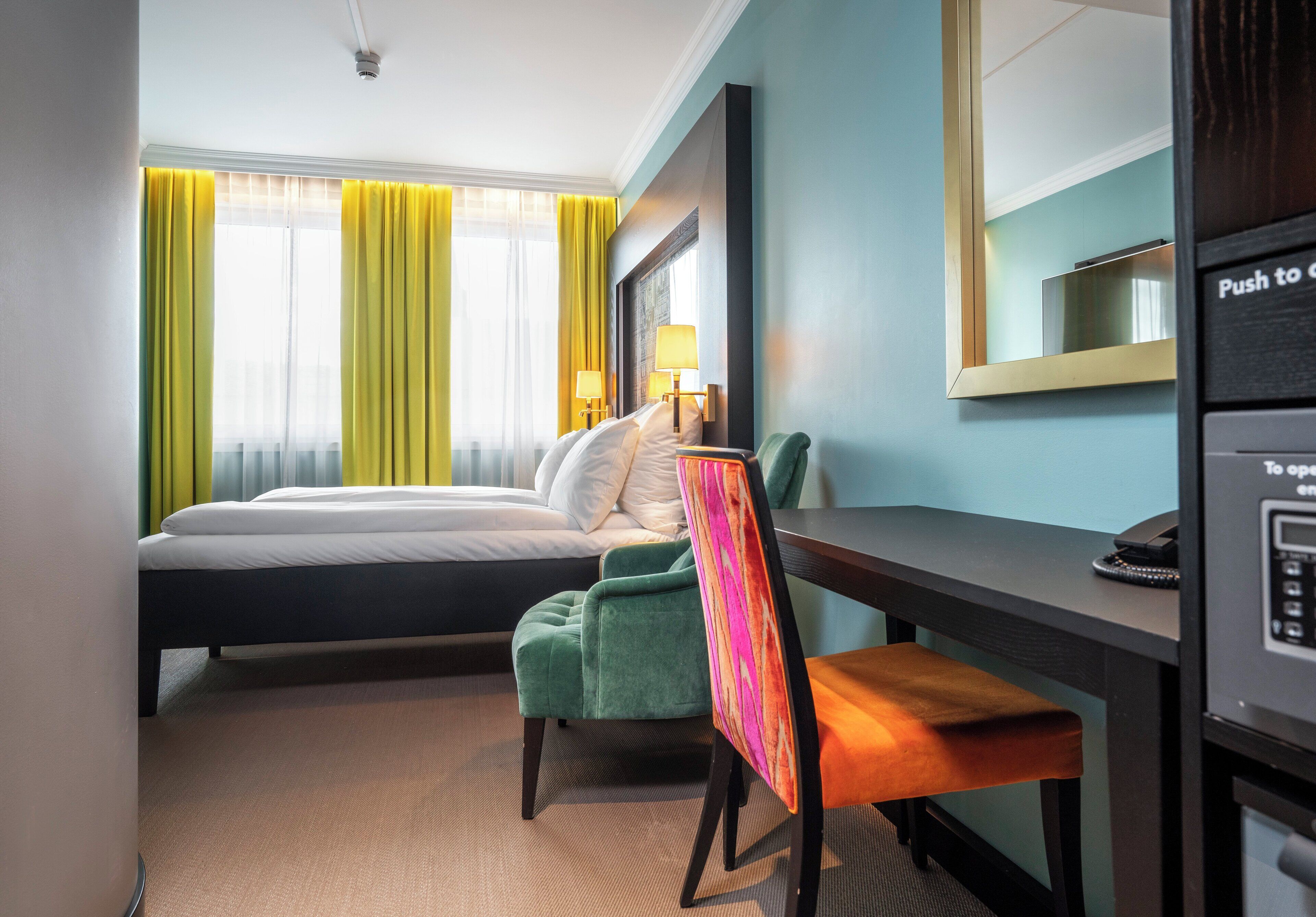 A hotel room with a bed, a desk, a chair, a mirror, and a telephone.
