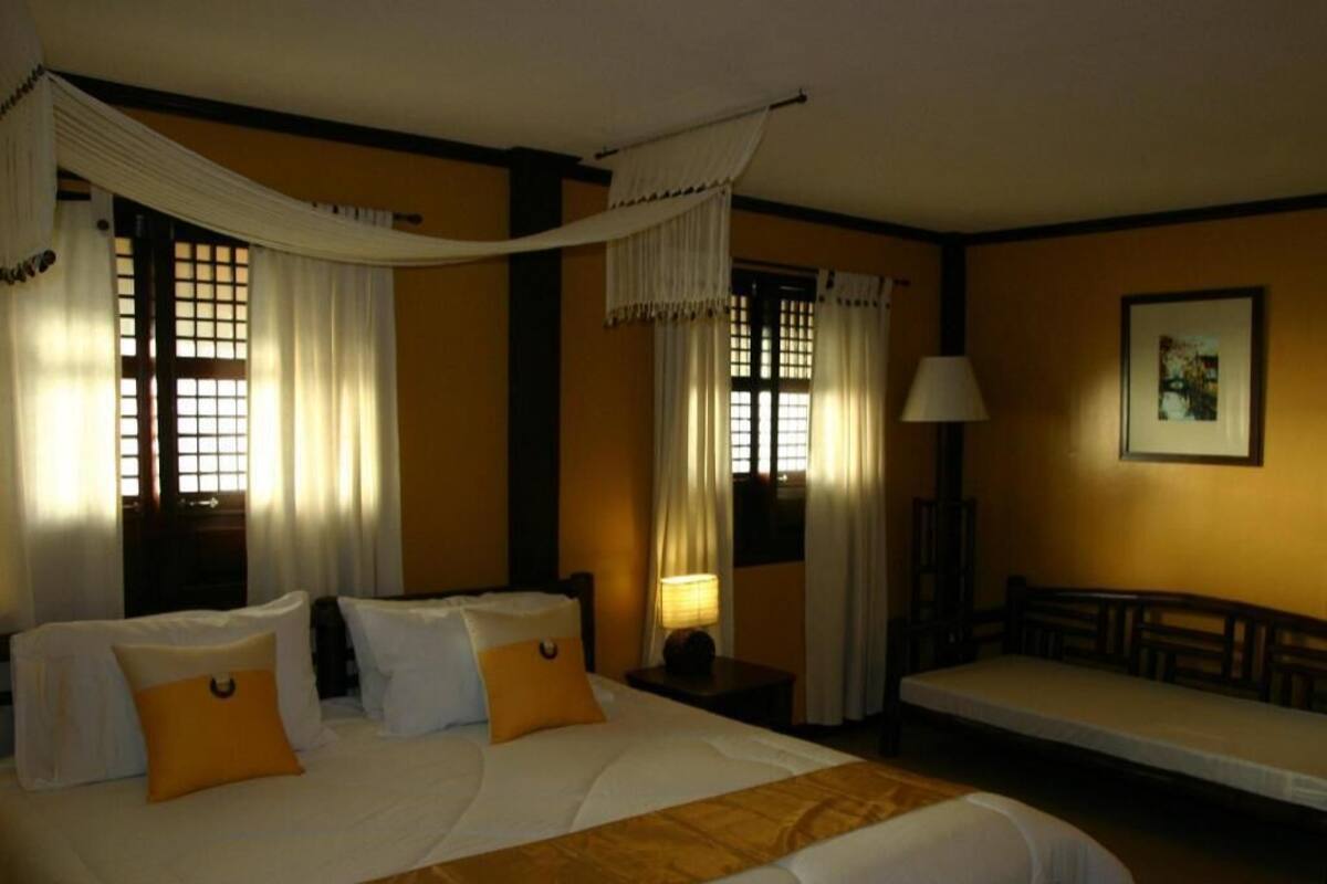 A hotel room with a large bed, two bedside tables, a bedside lamp, a sofa, and a window with curtains.