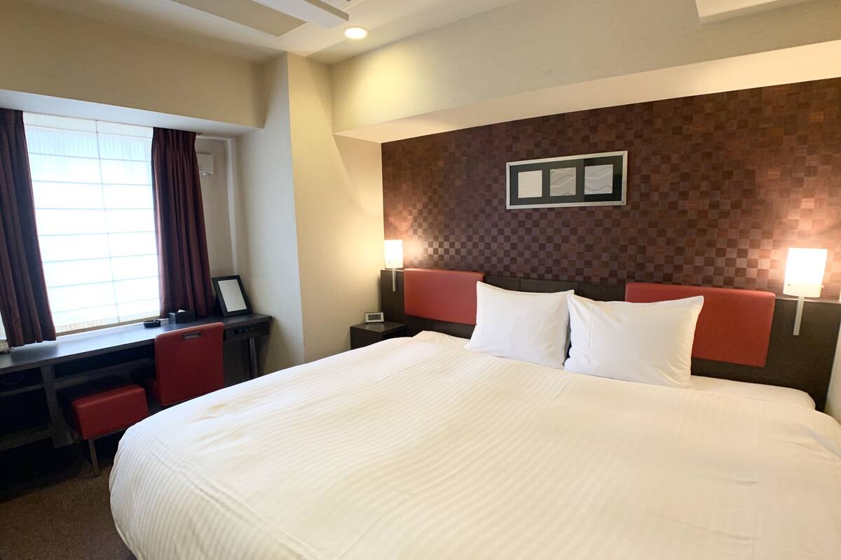 Una habitación de hotel con una cama grande, un escritorio con una silla, una ventana con cortinas y dos lámparas de noche.