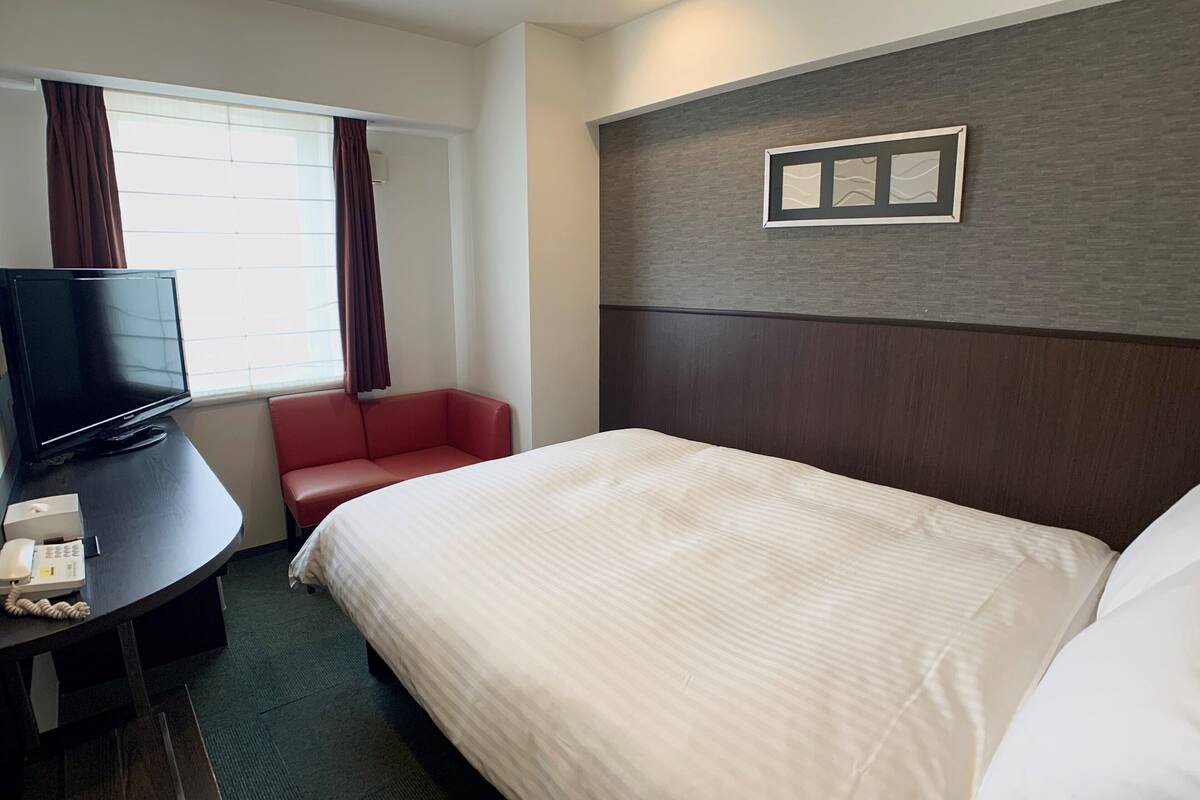 Habitación de hotel con una cama grande, un escritorio con televisor, una silla roja y una ventana con cortinas.