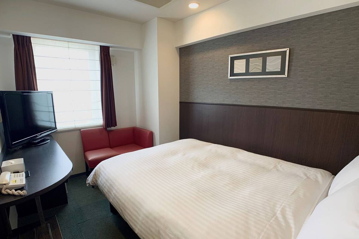 Habitación de hotel con cama, escritorio con teléfono, televisor, silla roja y ventana con cortinas.