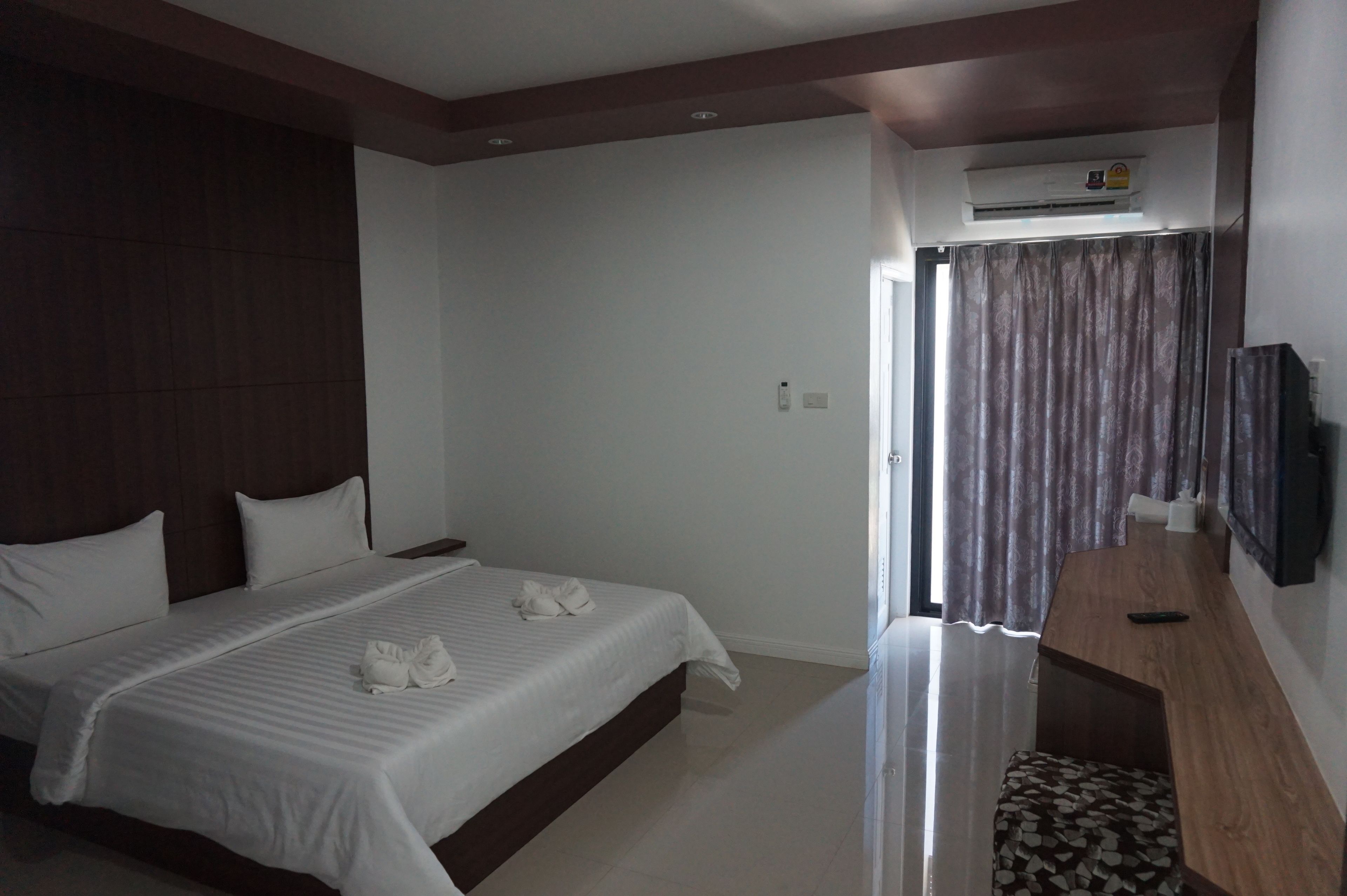 A hotel room with a large bed, a wooden headboard, a flat-screen TV mounted on the wall, and an air conditioning unit above the door.