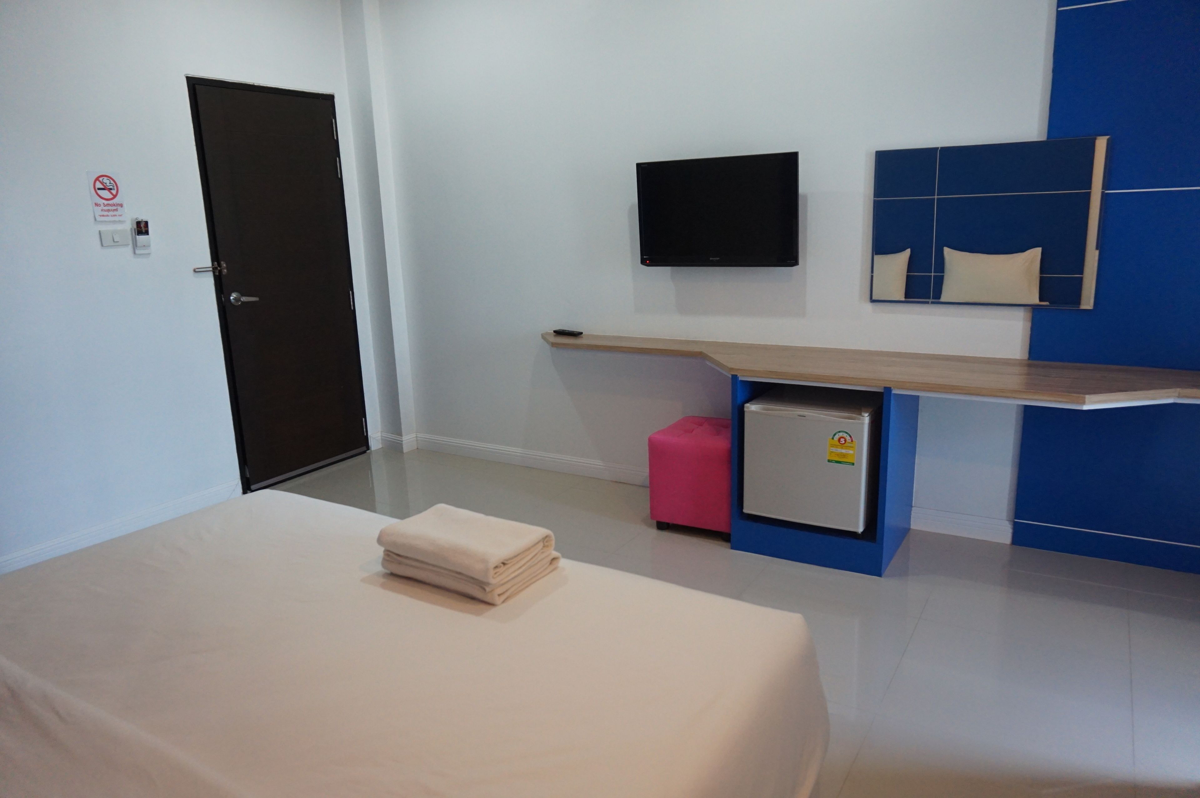 Habitación de hotel con cama, televisor montado en la pared, refrigerador pequeño, taburete rosa y estante de madera.