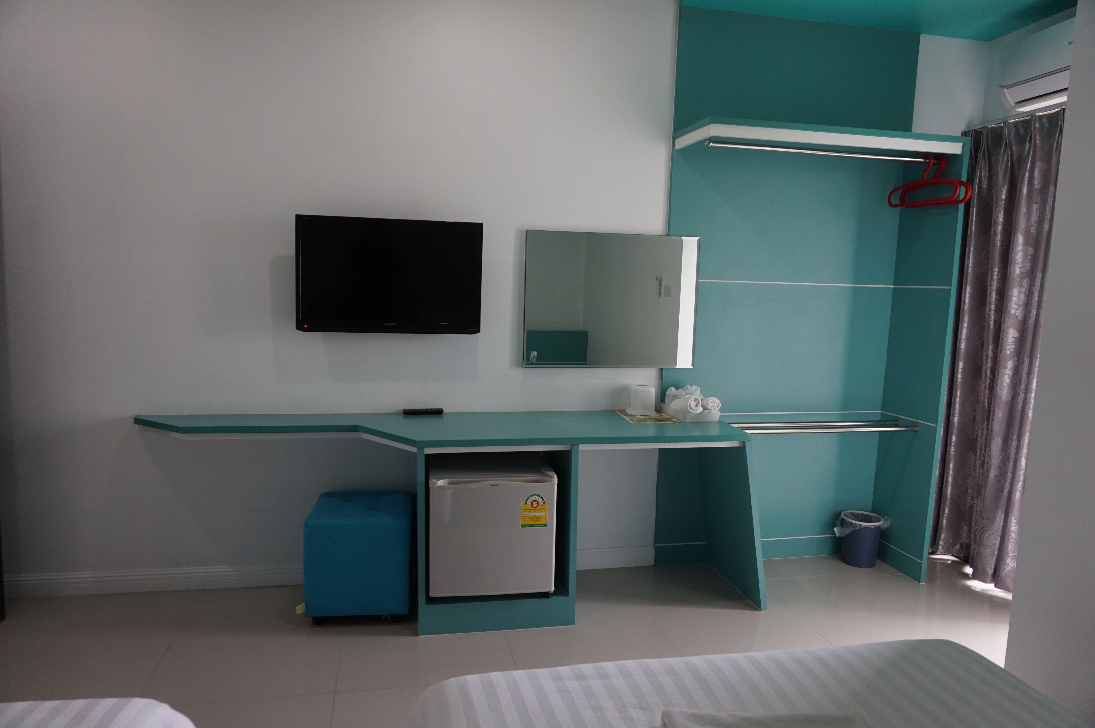 A modern hotel room with a teal-colored desk, a small refrigerator, a flat-screen TV mounted on the wall, a mirror, and a shower area with a curtain.