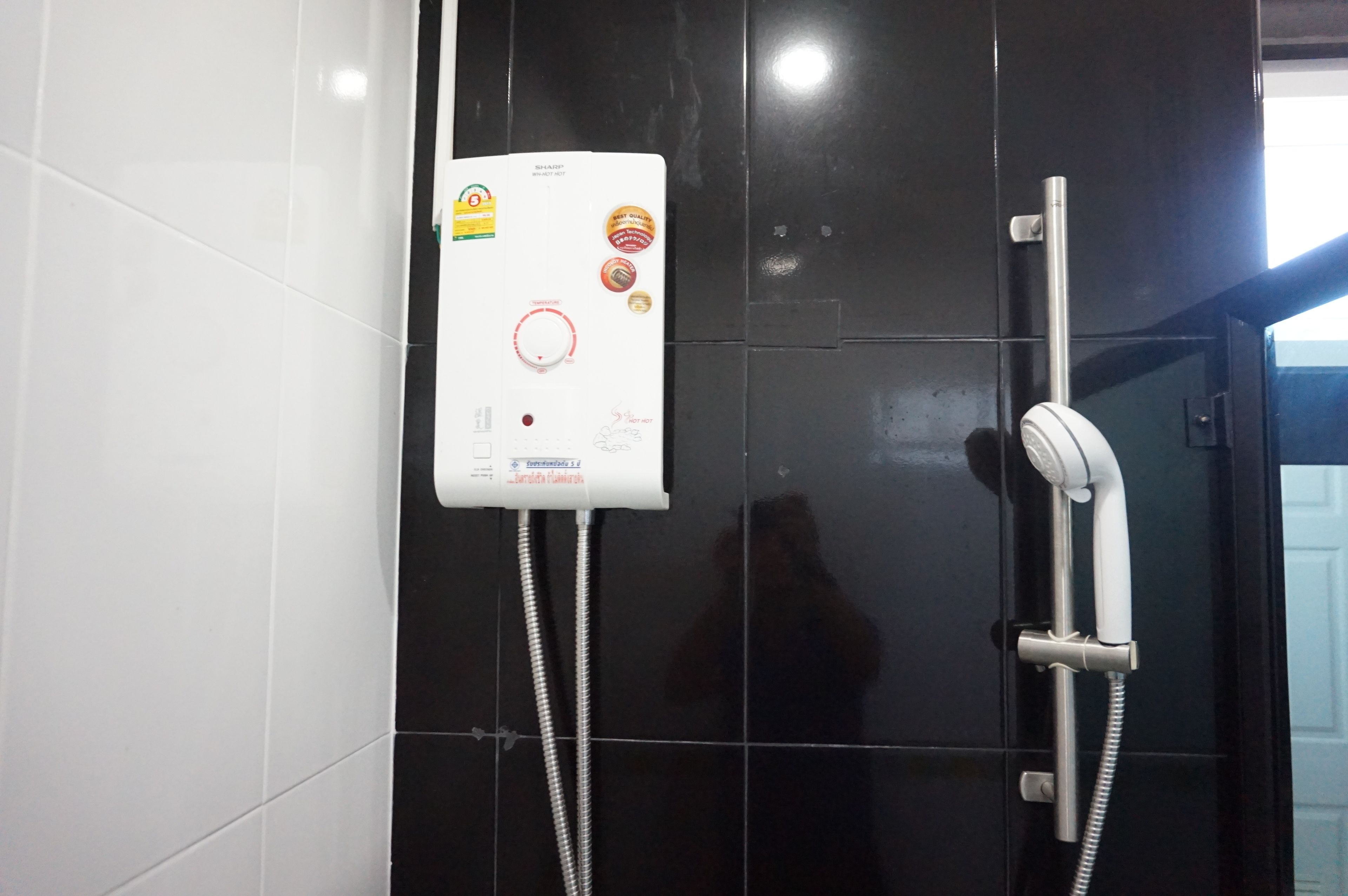 A bathroom with a white electric shower unit mounted on a black tiled wall, a handheld showerhead, and a control panel with various stickers and labels.