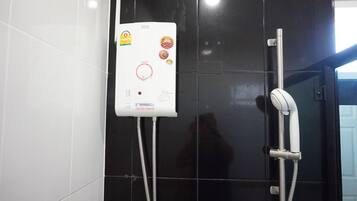 A bathroom with a white electric shower unit mounted on a black tiled wall, a handheld showerhead, and a control panel with various stickers and labels.