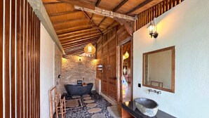 A rustic bathroom with a stone wall, wooden beams, a freestanding bathtub, a stone basin, and a wooden door. - Royal Casa Ganesha (Mas)