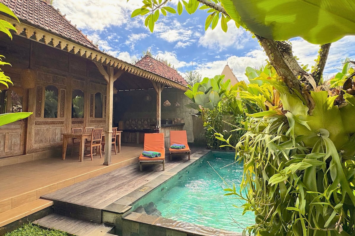 A poolside area with a wooden deck, lounge chairs, and a covered seating area with a tiled roof.