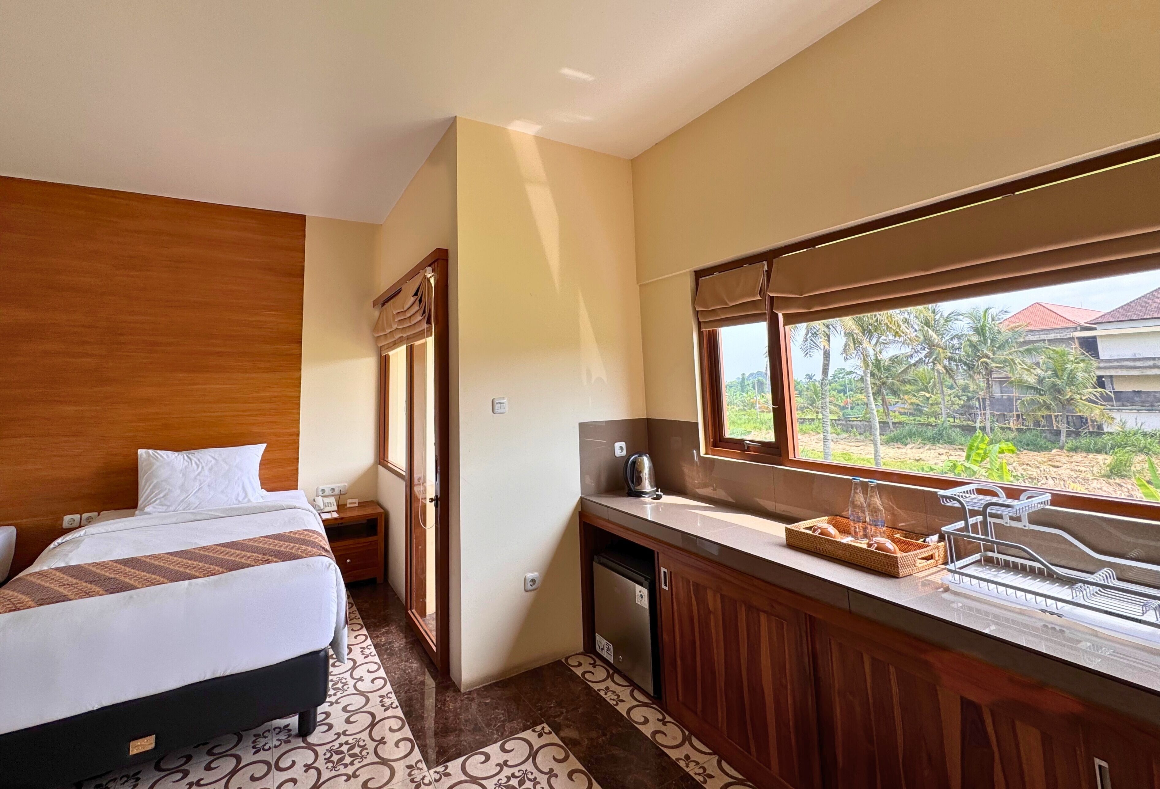 Habitación de hotel con cama, una kitchenette pequeña y ventana que ofrece vista a palmeras y un edificio.