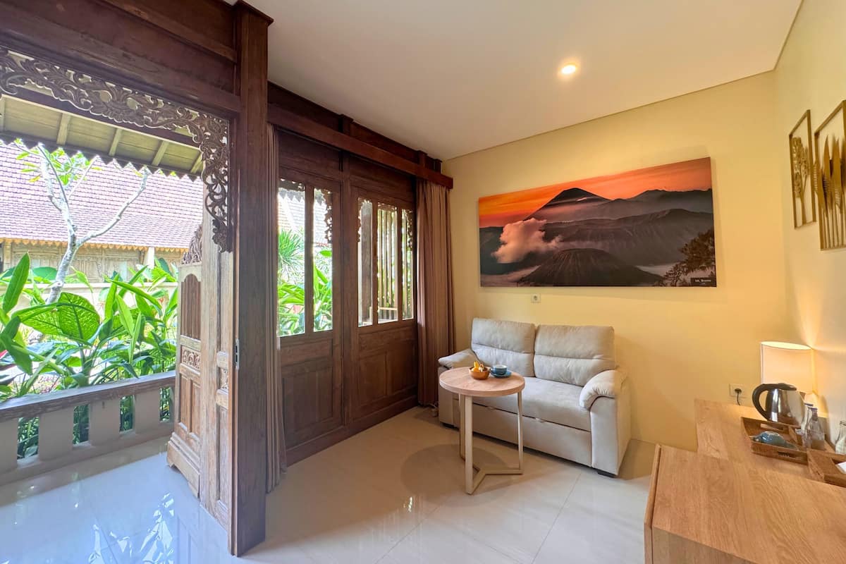 A room with a sofa, a small table, a painting of a sunset, and a view of greenery outside.