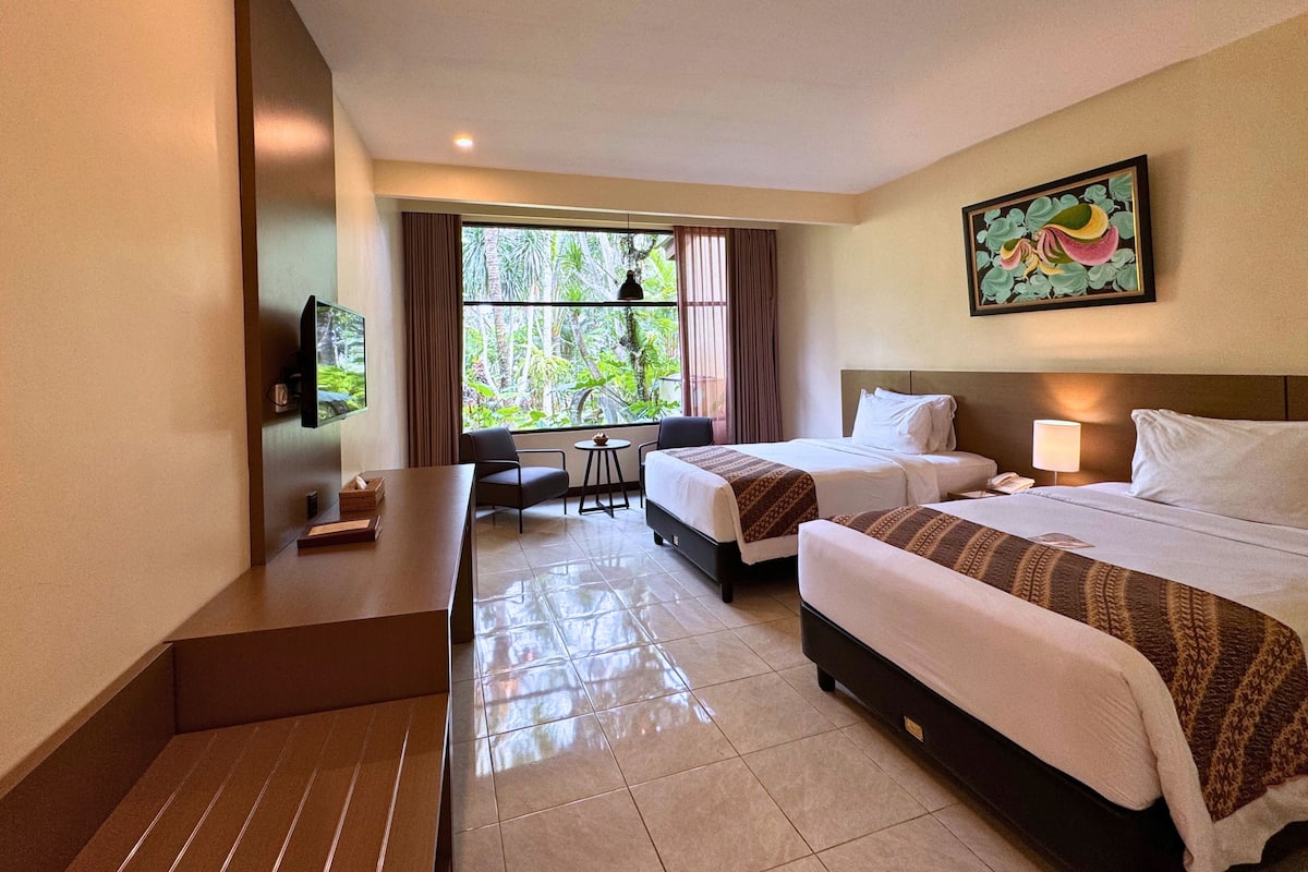 A hotel room with two beds, a desk, a chair, and a window with a view of greenery.