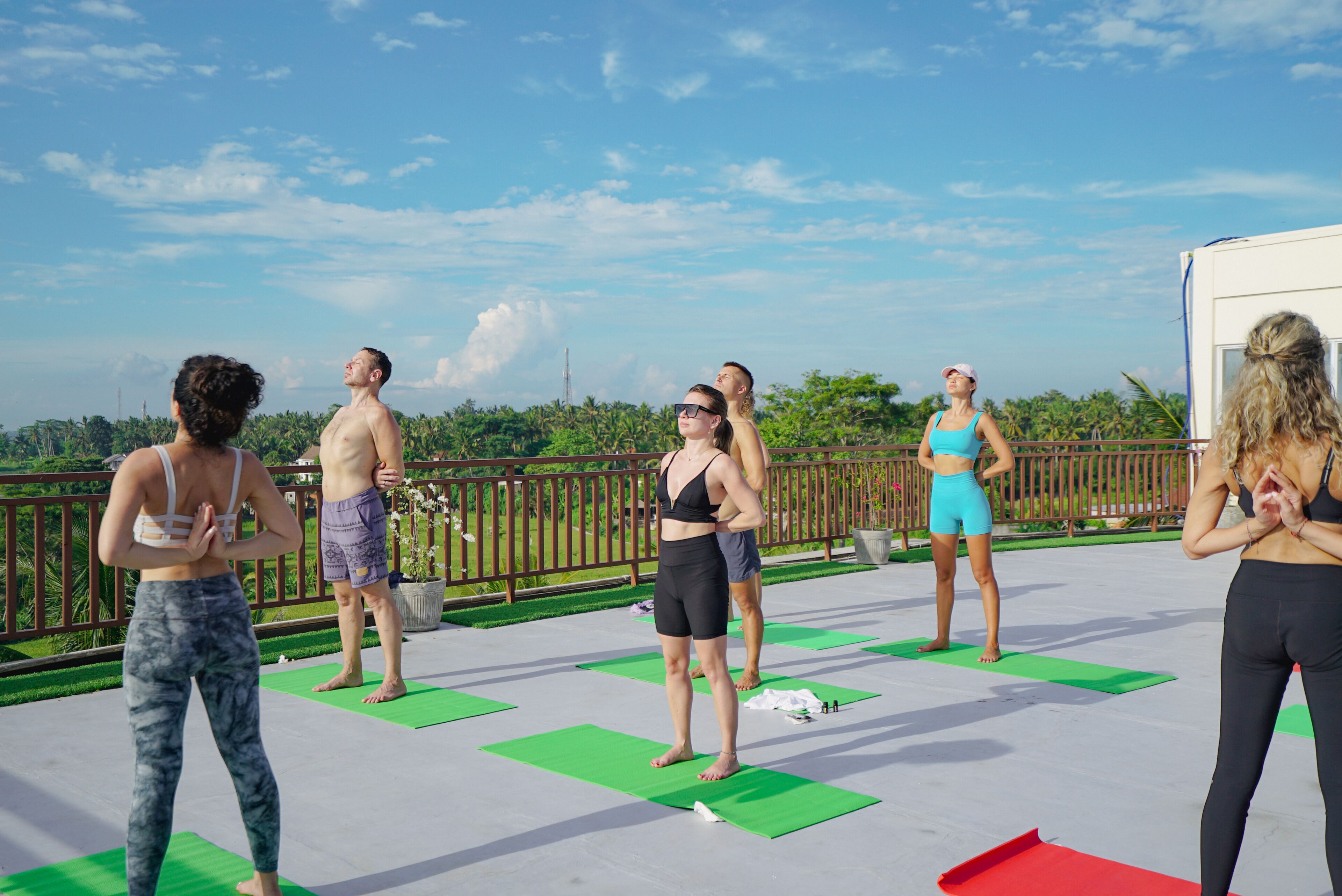 A group of people on a rooftop deck with green exercise mats, a railing, and a clear sky.