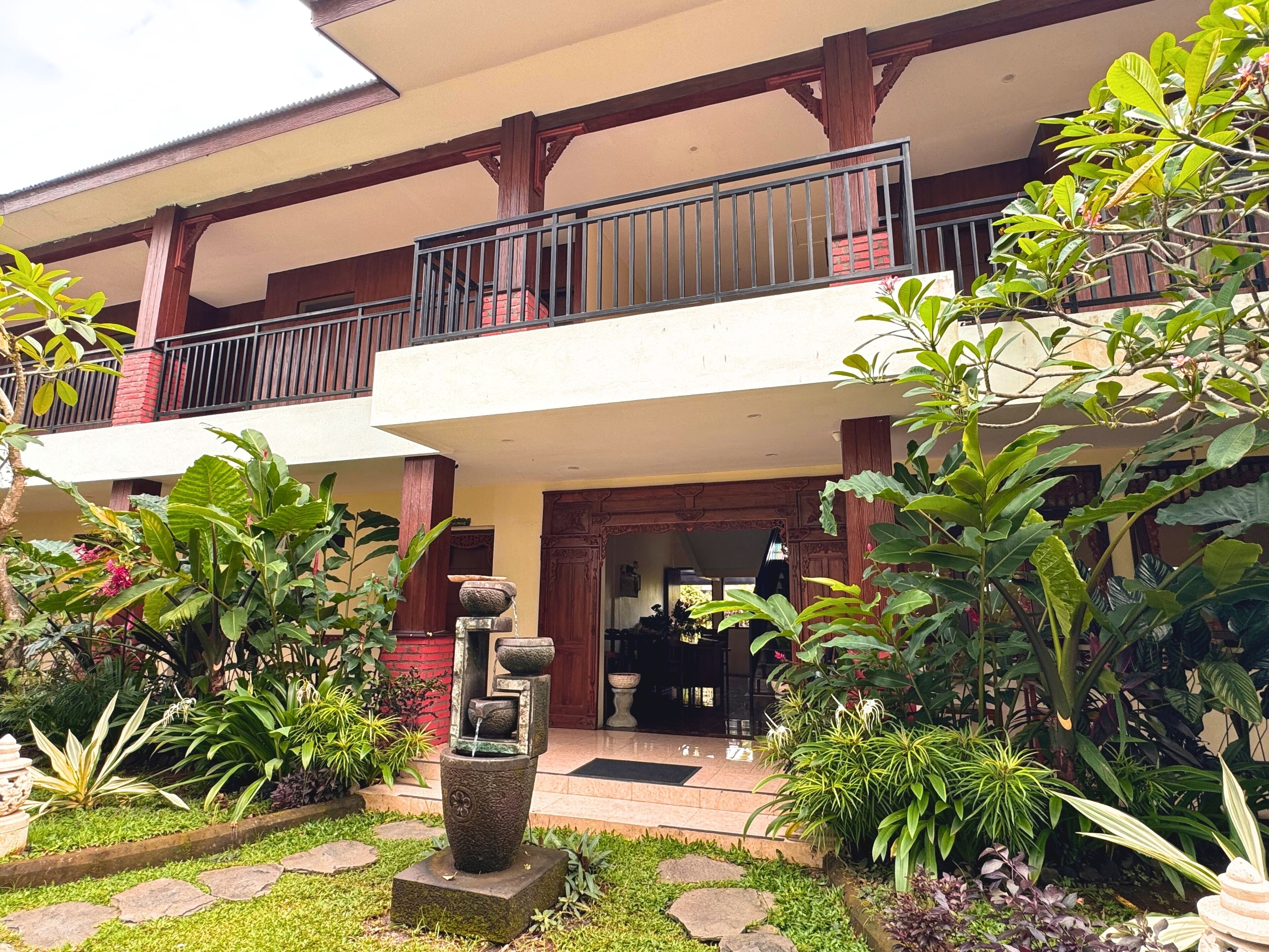 A two-story building with a balcony, a stone water feature, and lush greenery.