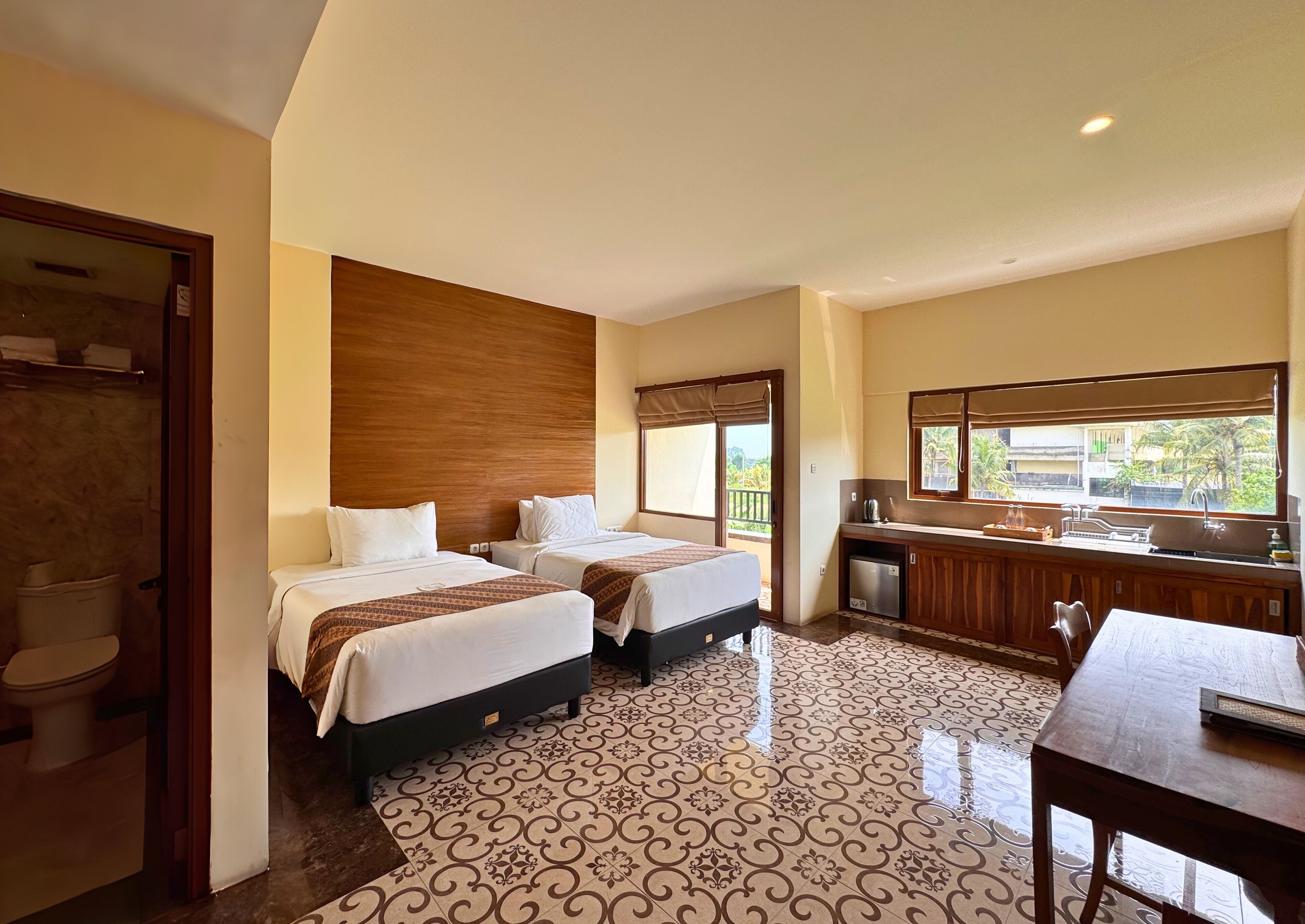 A hotel room with two beds, a small kitchenette, a bathroom, and a view of greenery.