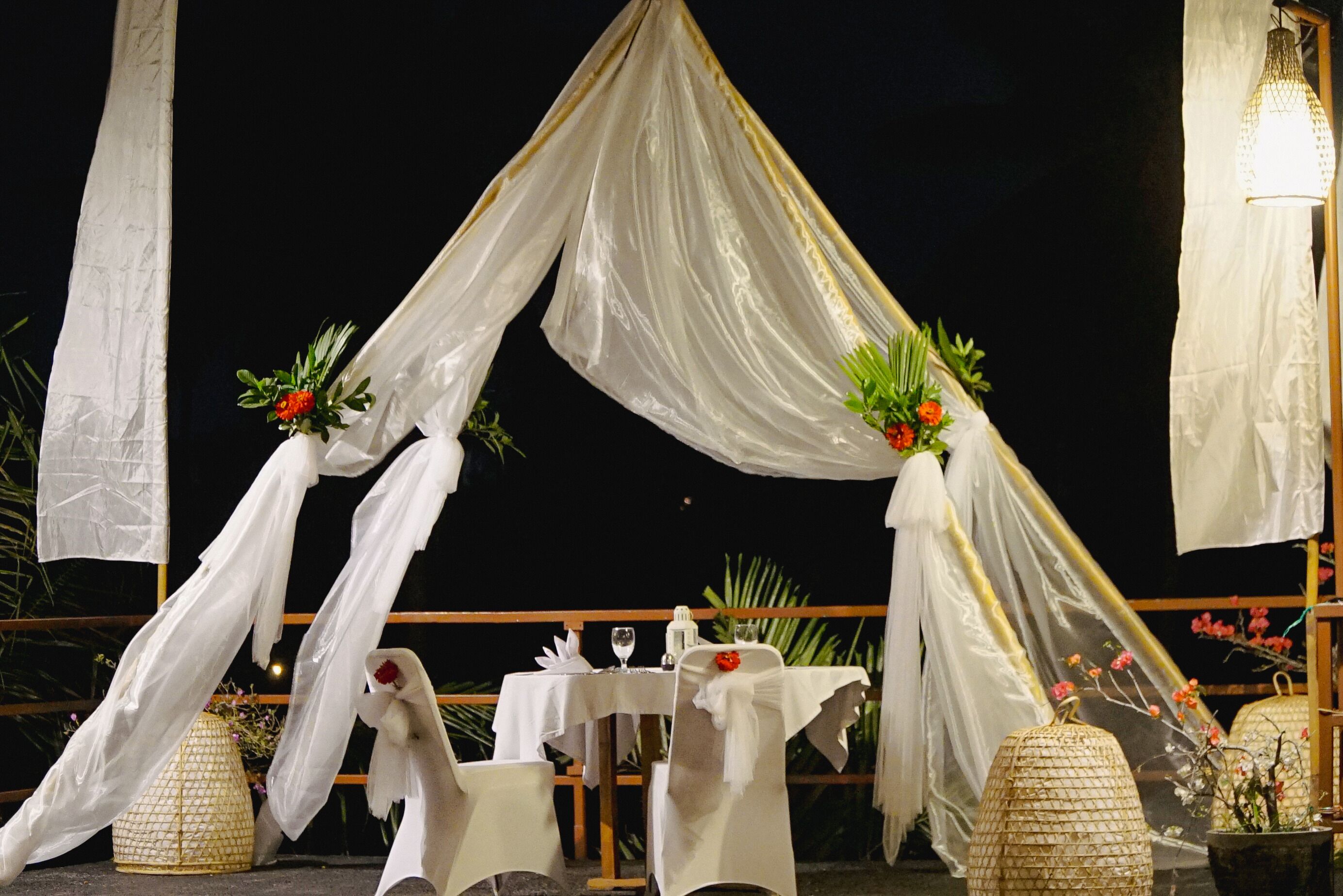 Un espacio de comedor al aire libre con mesas y sillas blancas, decoraciones de tela drapeada y plantas en macetas.