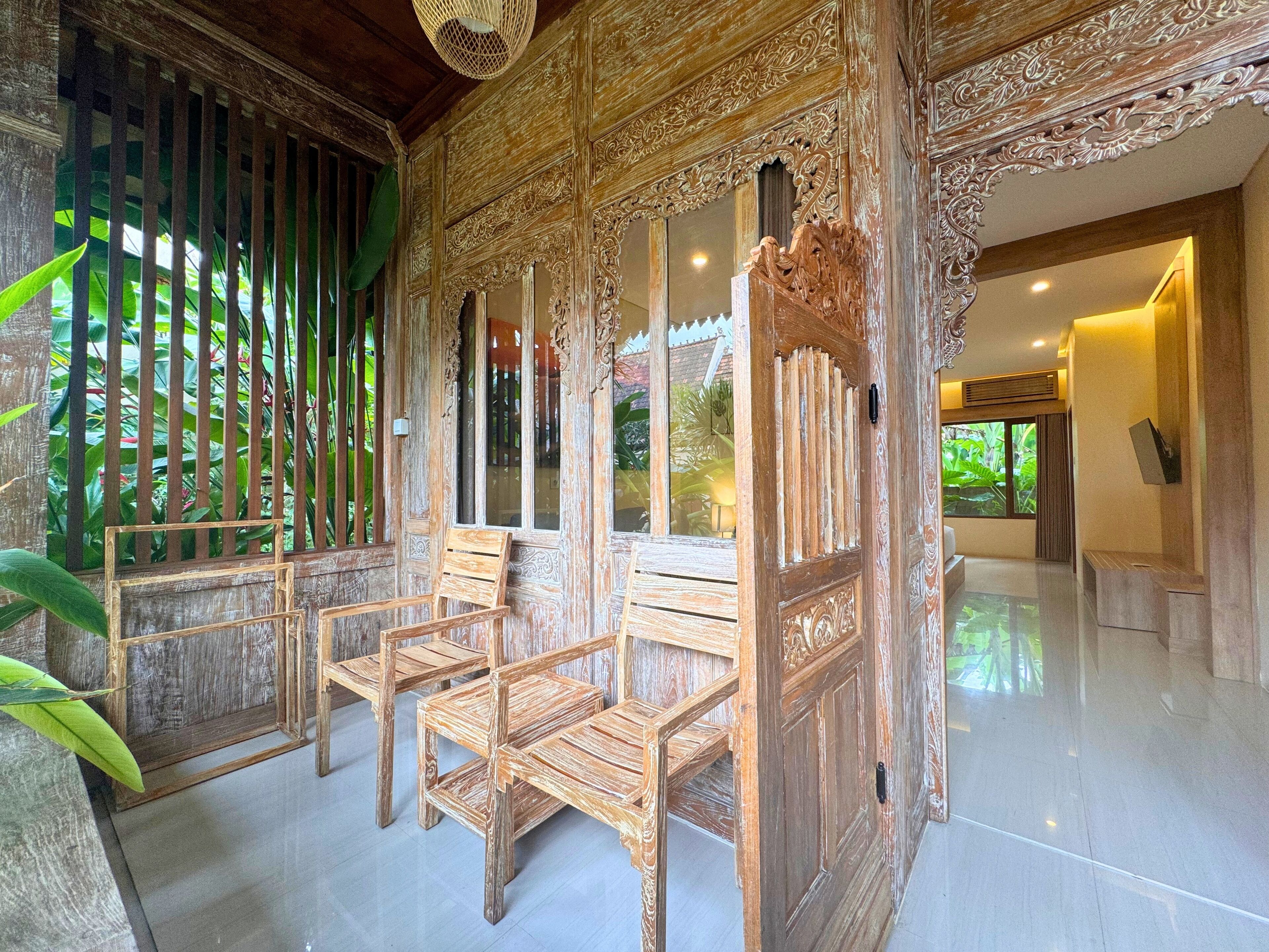 A traditional wooden room with ornate carvings, a tiled floor, and a view of greenery outside.