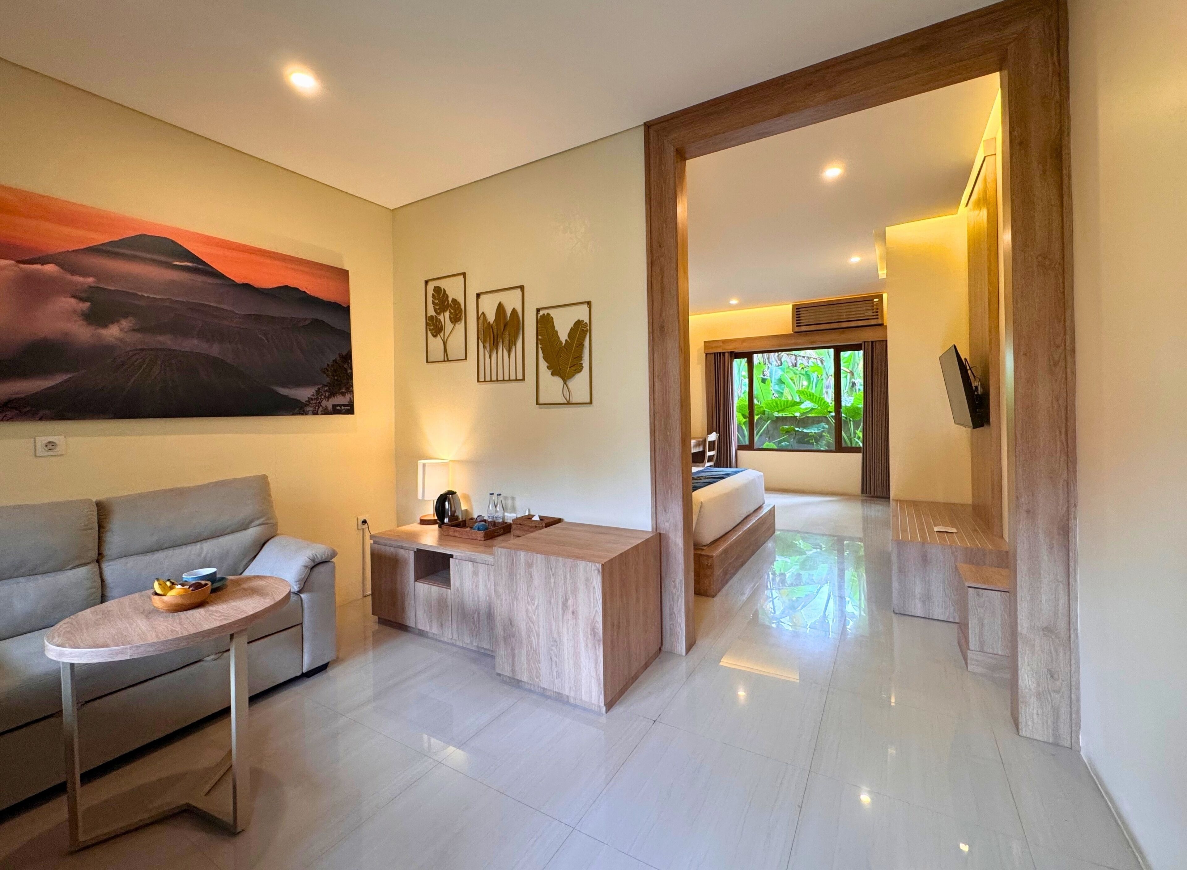 A modern hotel room with a large painting, a wooden desk, a sofa, and a bed with a view of greenery outside.