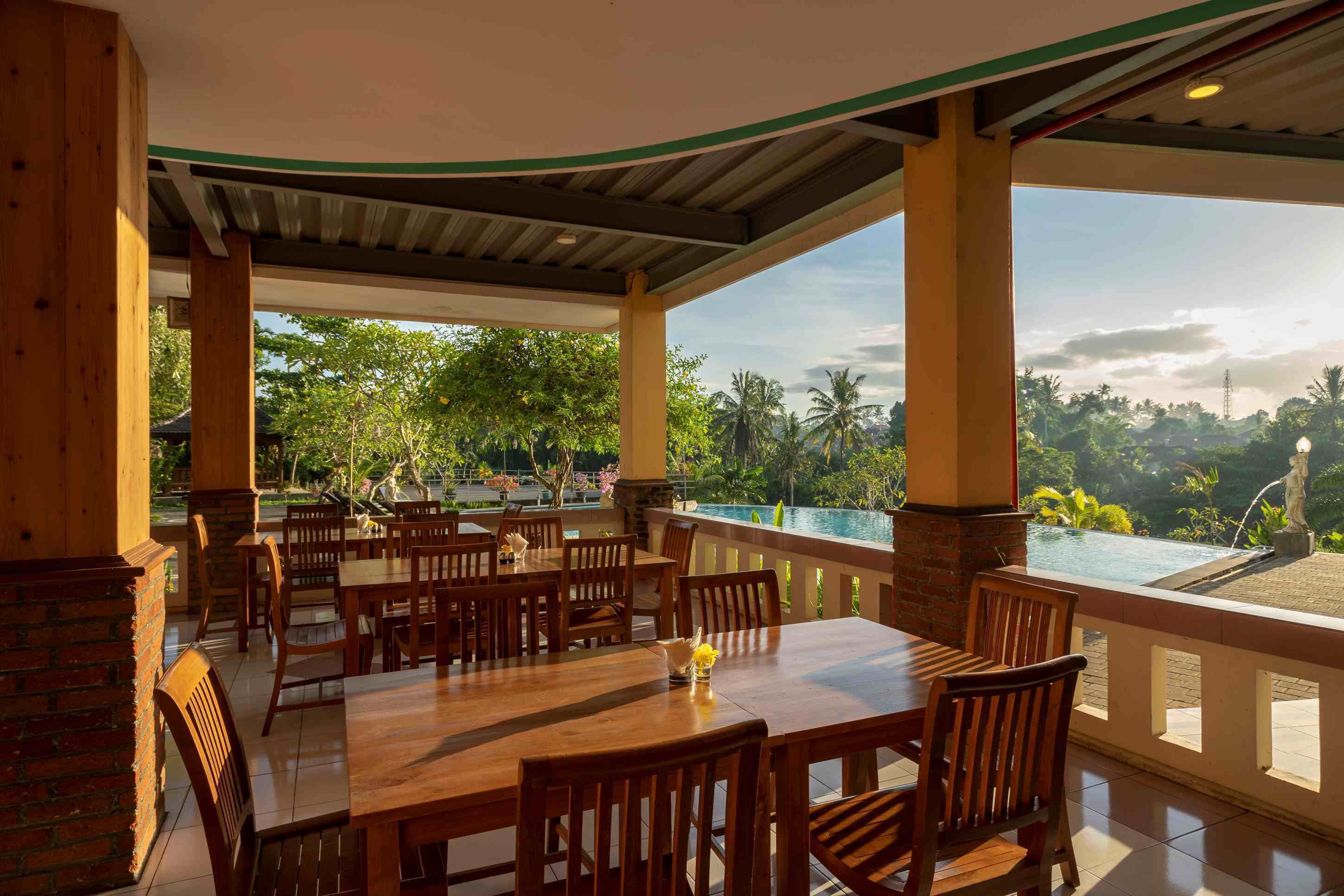 A covered outdoor dining area with wooden tables and chairs, overlooking a pool and lush greenery.