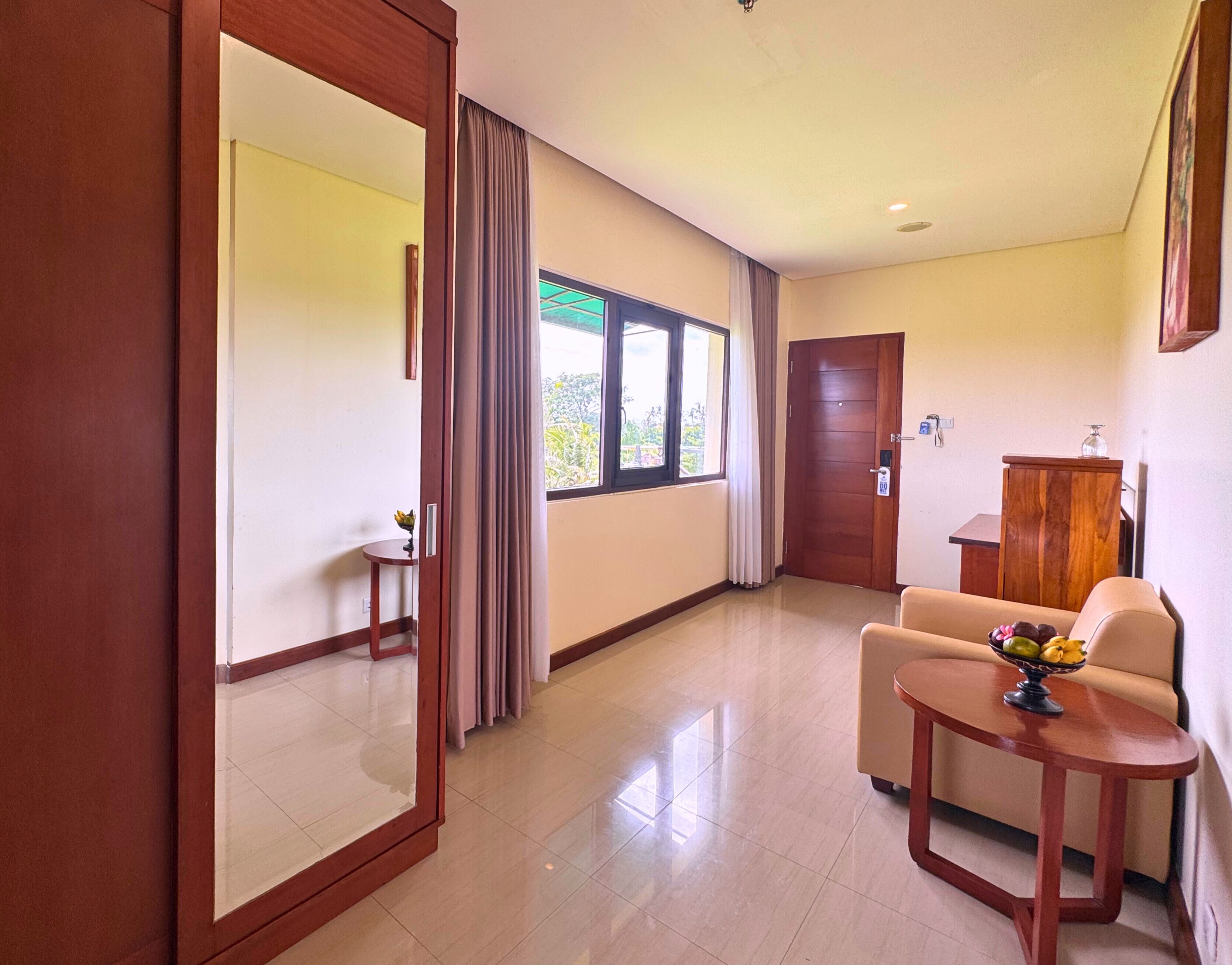 A hotel room with a large mirror, a wooden door, a window with curtains, a sofa, a small table with fruit, and a wooden cabinet.