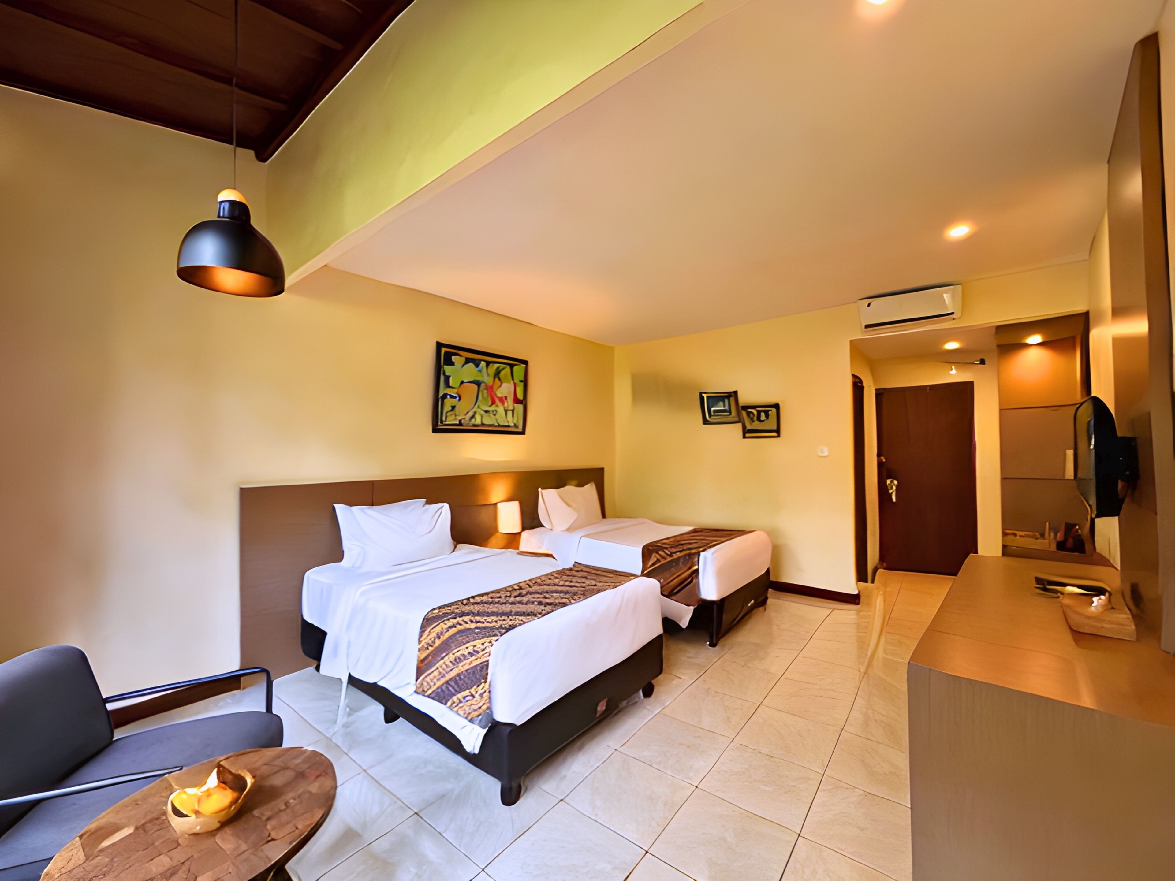 A hotel room with two beds, a desk, a chair, a small table with a candle, and a painting on the wall.