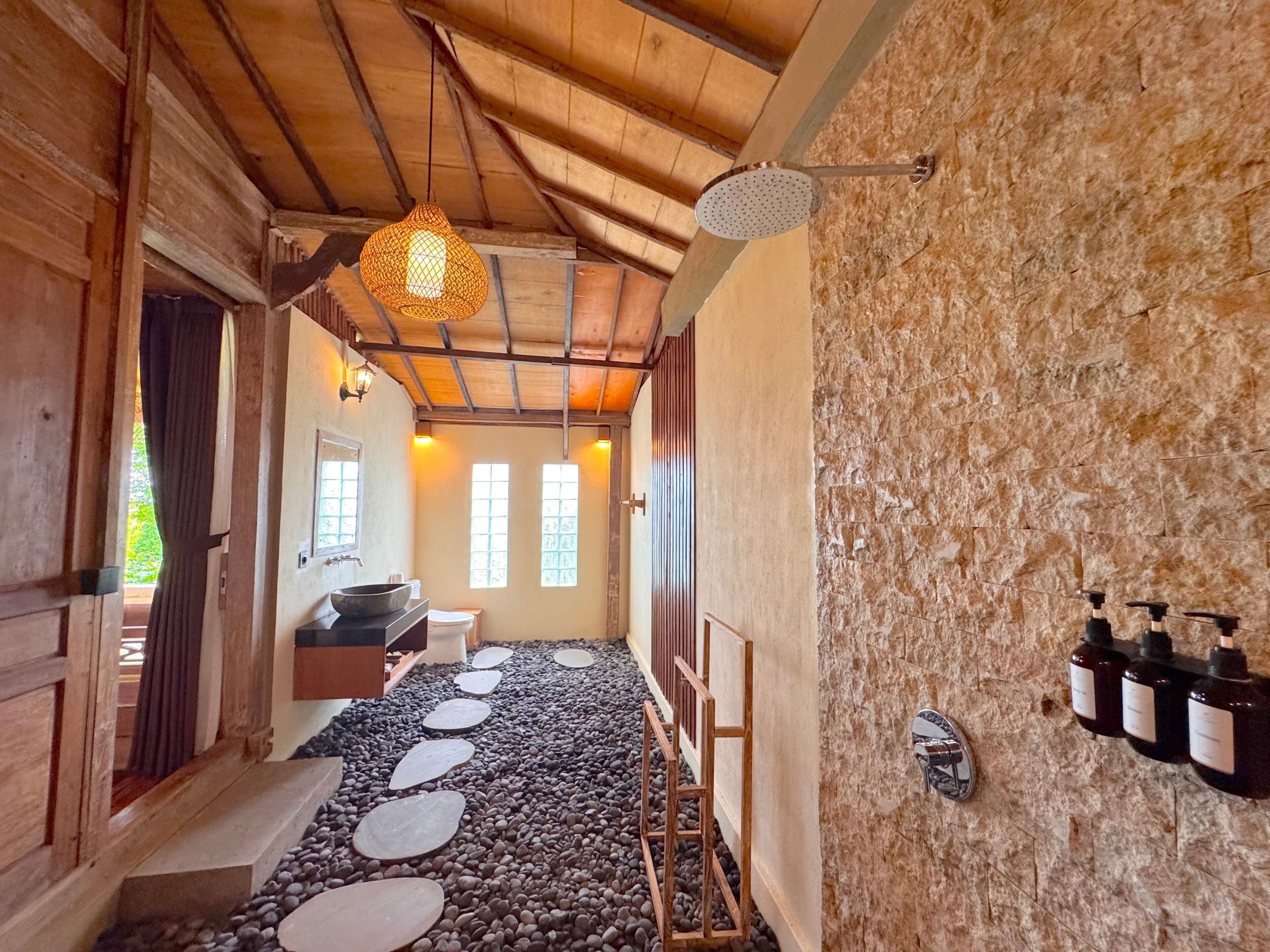 A rustic bathroom with a stone wall, wooden beams, a stone pathway, and a hanging light fixture.