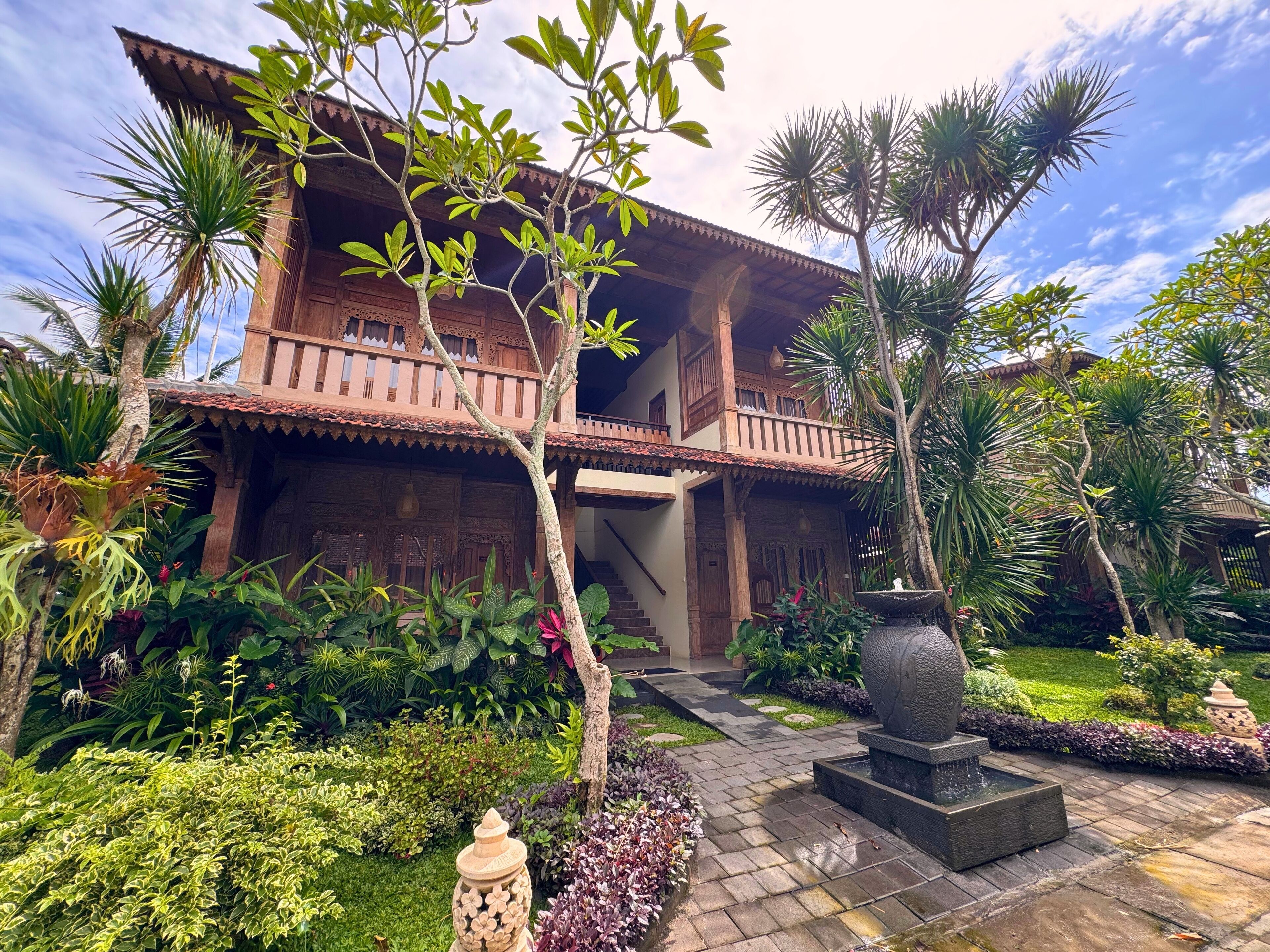 A traditional wooden house with a balcony, surrounded by lush tropical plants and a stone fountain.