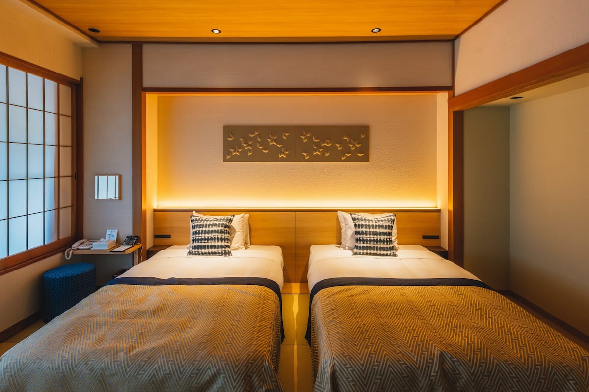A hotel room with two beds, a wooden ceiling, a wall-mounted phone, and a window with a view.