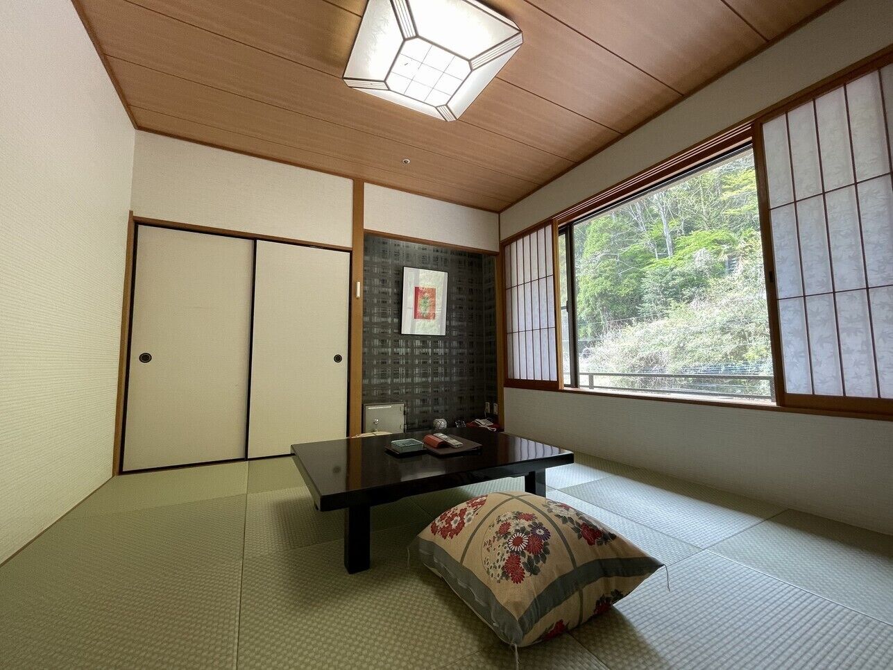 A traditional Japanese room with tatami flooring, a low table, a cushion, sliding doors, and a window with a view of greenery.