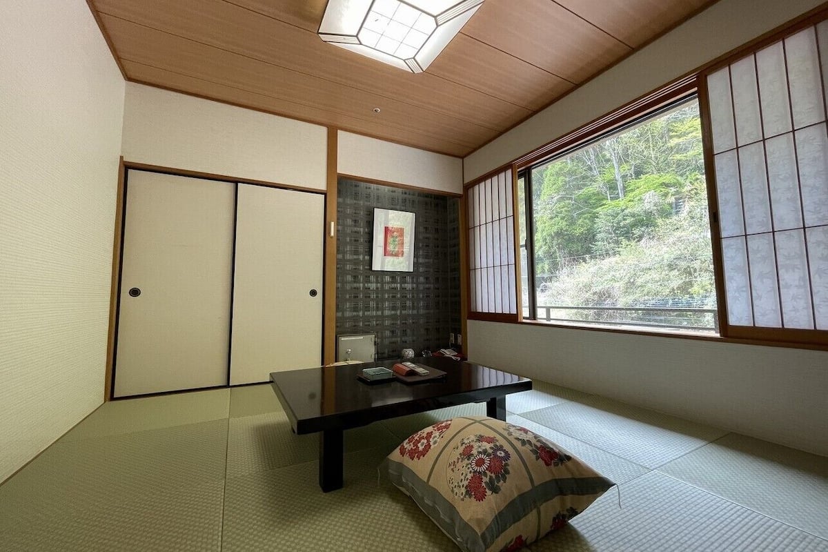 A traditional Japanese room with tatami flooring, a low table, a cushion, sliding doors, and a window with a view of greenery.