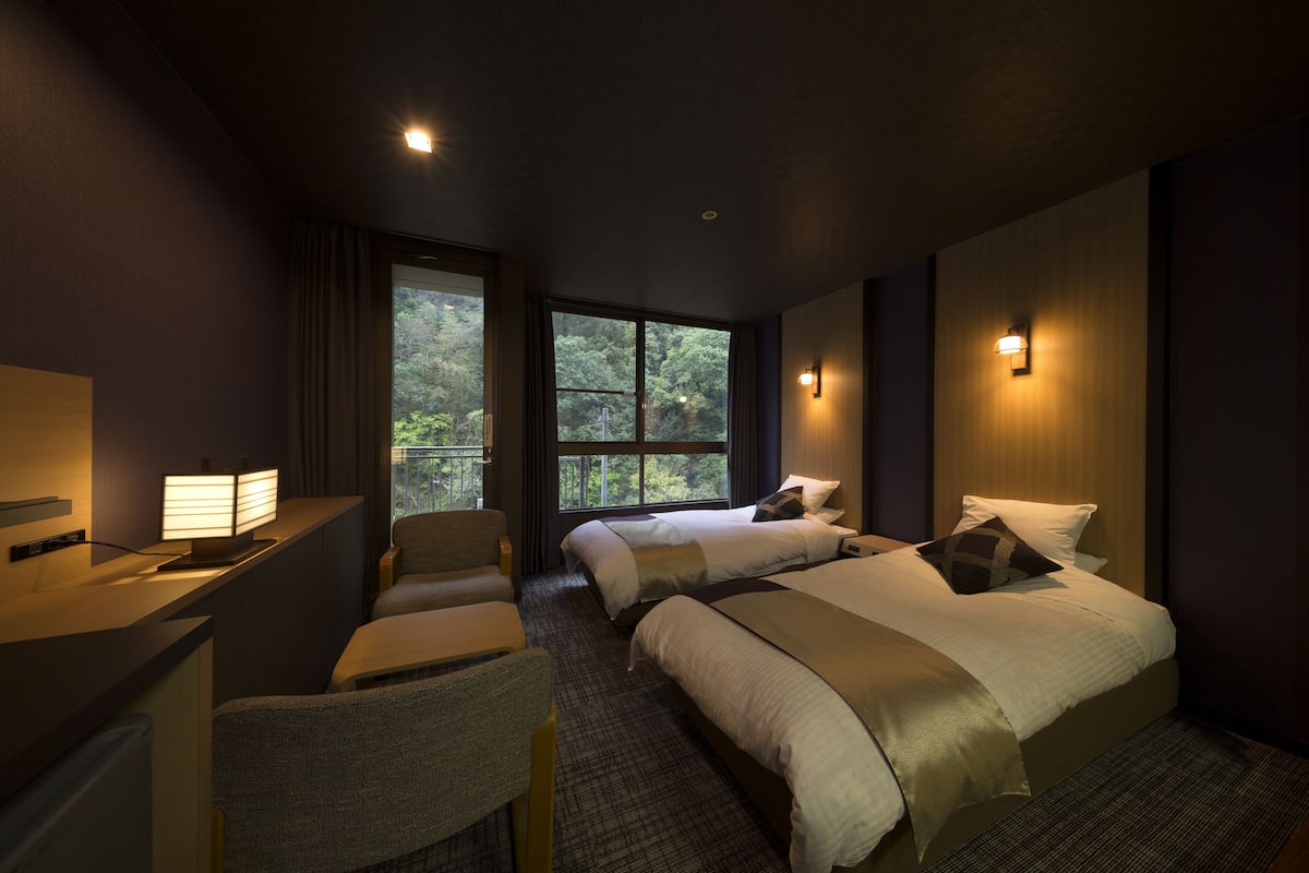 A hotel room with two beds, a desk, and a chair. There are windows providing a view of trees.