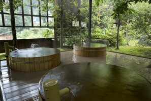 Perinteinen japanilainen onsen-kylpylä, jossa on puiset kylpyaltaat ja kivinen lattia. Kylpylän ympärillä on vehreää kasvillisuutta ja se on aidattu puuaidalla.