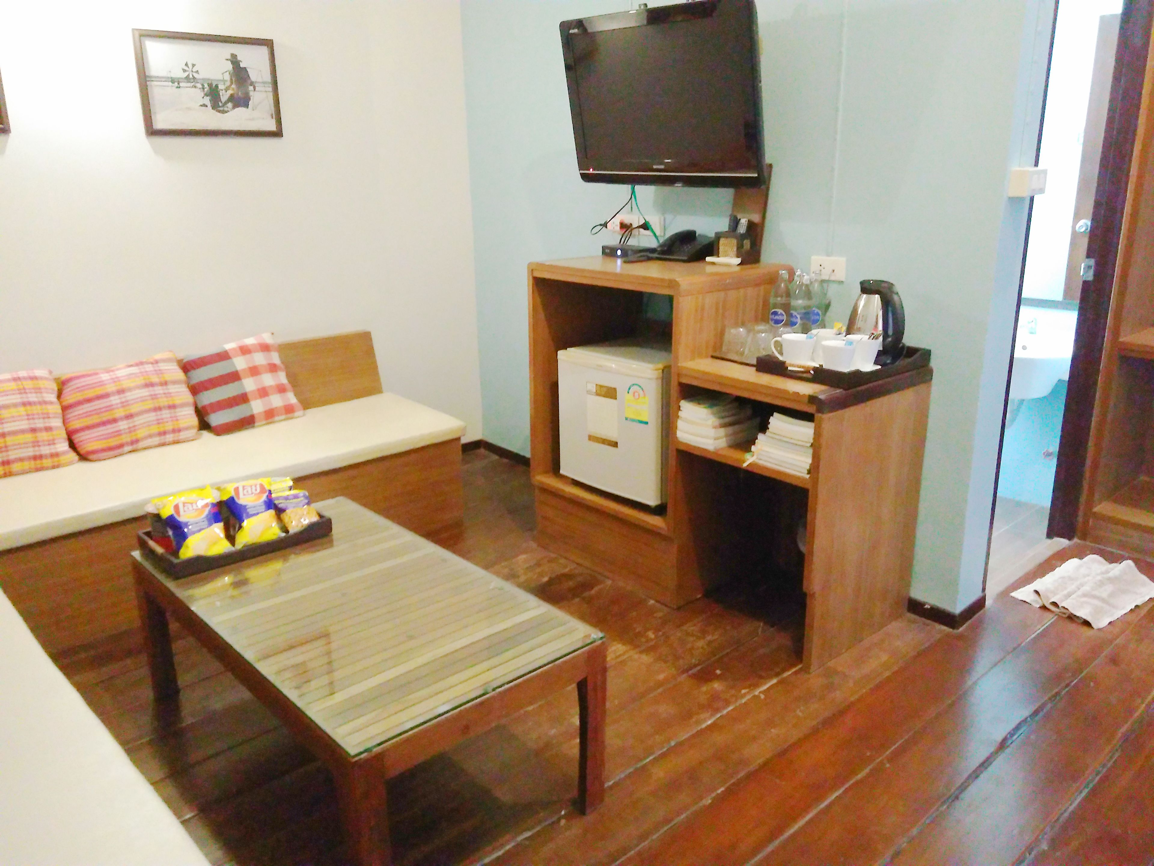 Una sala de estar con una mesa de centro de madera, un sofá blanco con cojines a rayas, un televisor montado en la pared y un refrigerador pequeño.