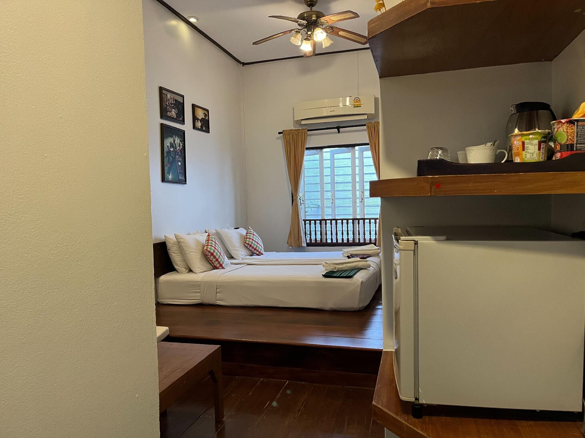 Habitación de hotel pequeña con cama, una mesita, ventilador de techo y refrigerador.