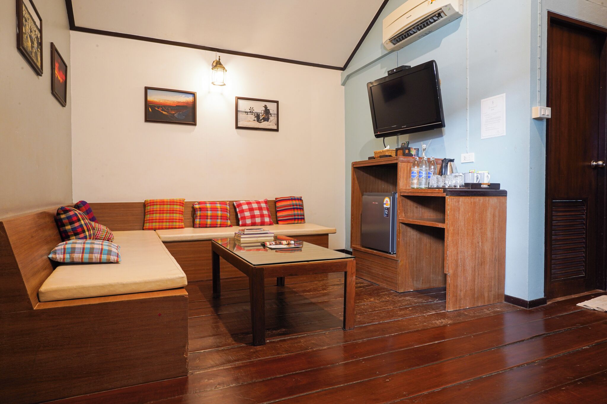 A living room with a sofa, a coffee table, a television, and a small kitchen area with a refrigerator and a shelf with bottles and cups.
