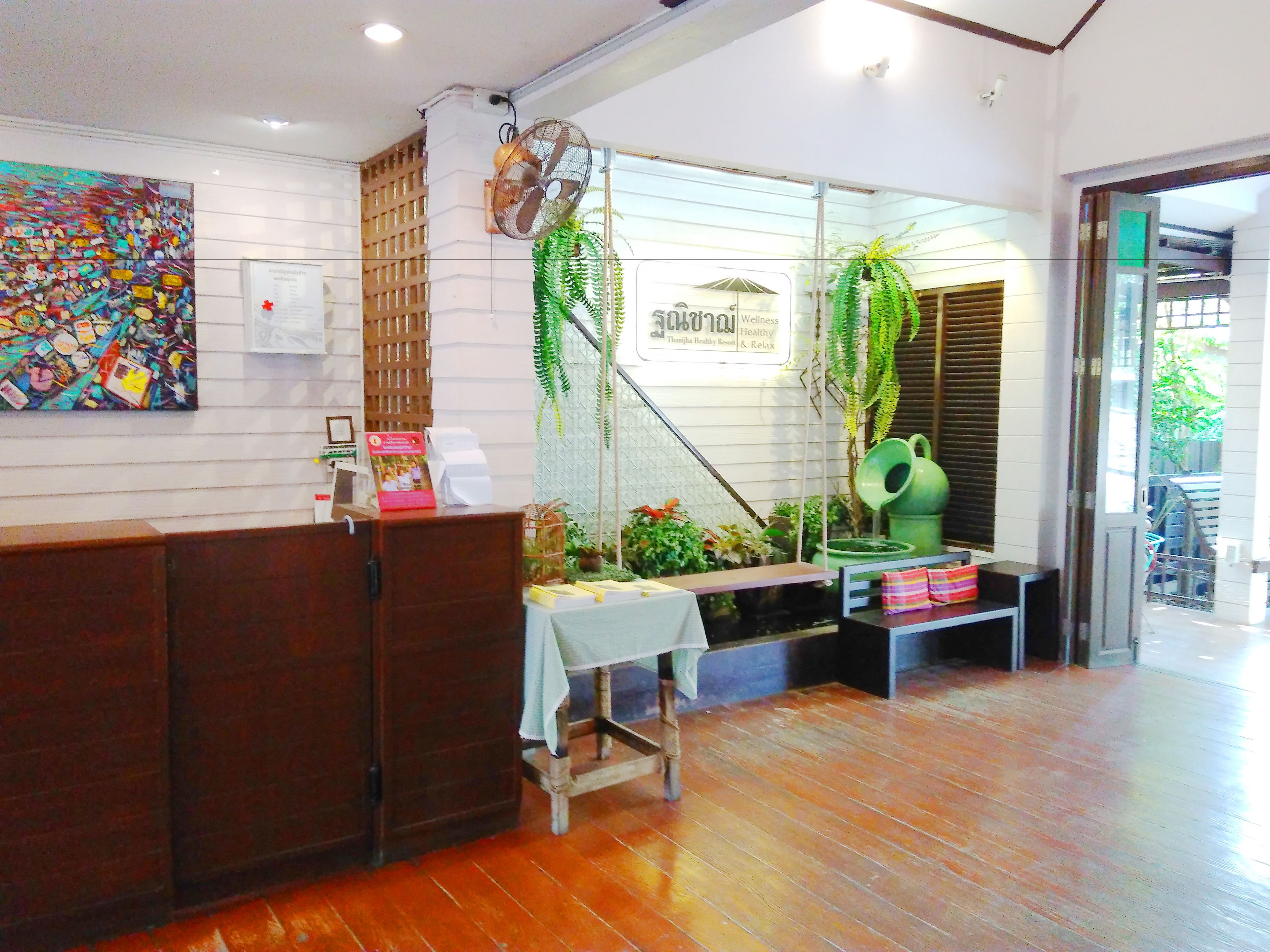 Un área de recepción con un mostrador de madera, una mesita con mantel blanco y un cartel que dice “Wellness & Relax”. Hay plantas en macetas y una regadera verde sobre la mesa.