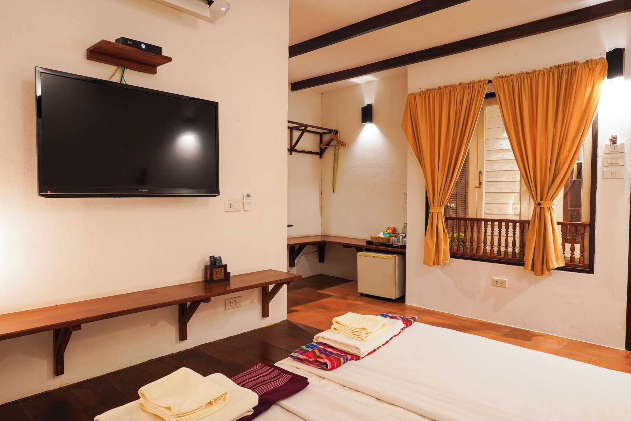 A hotel room with a flat-screen TV mounted on the wall, a wooden bench, a bed with neatly folded towels, and a window with curtains.