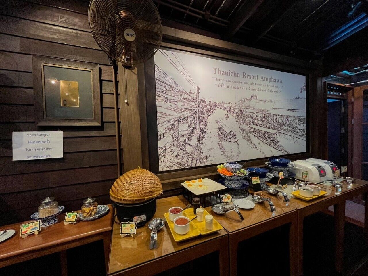 A buffet setup with various dishes and condiments, a fan, and a large map of Thanicha Resort Amphawa.