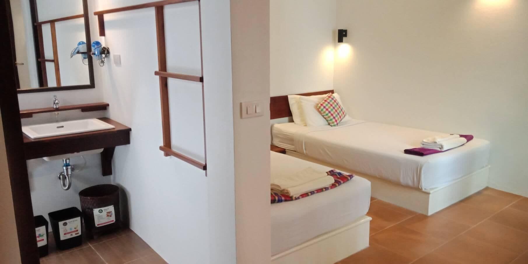 Ein Badezimmer mit Waschbecken, Spiegel und Mülleimern. Ein Bett mit Kopfkissen und Decke.