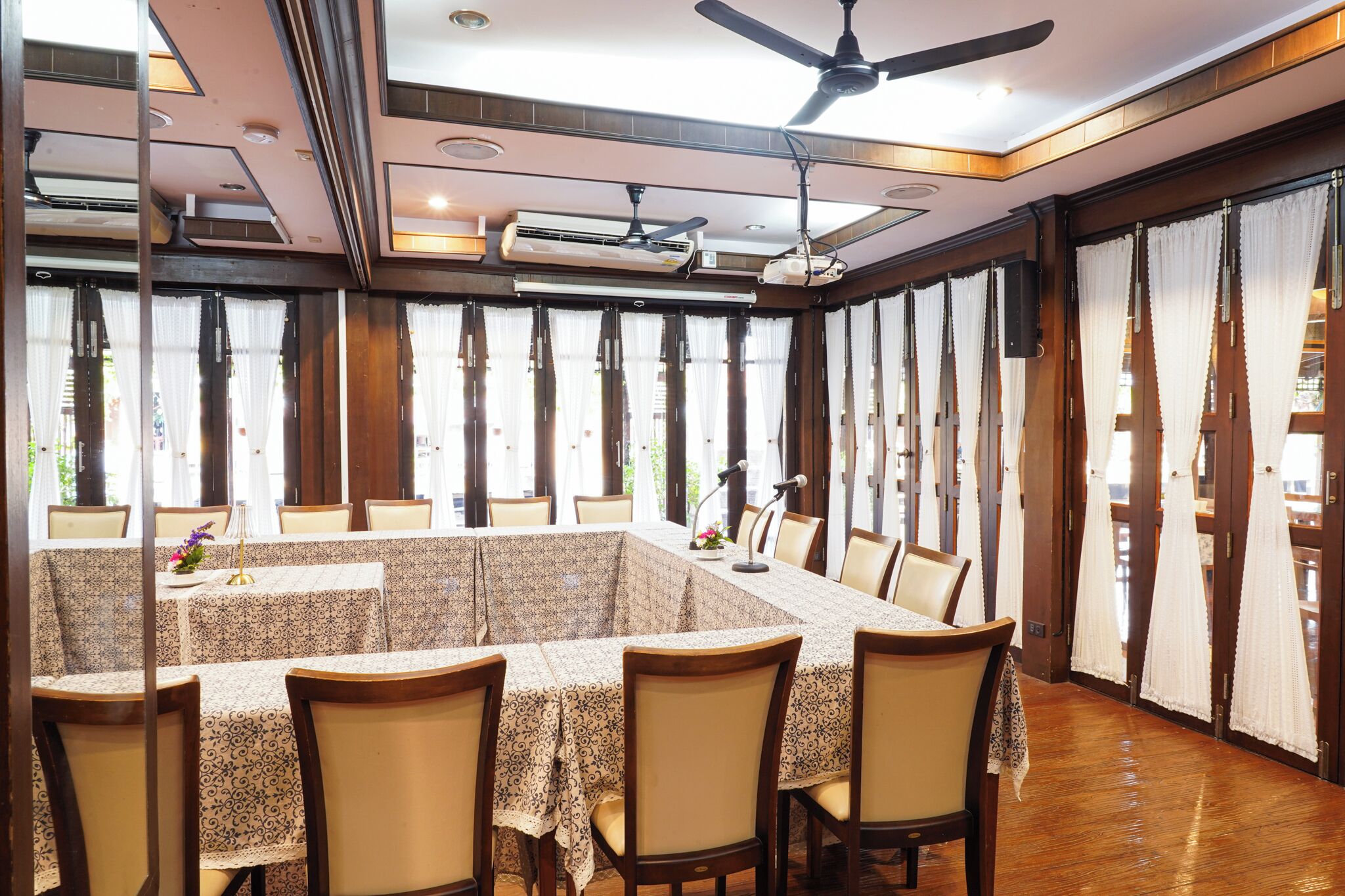Un comedor con una mesa grande, sillas, ventiladores de techo y ventanas amplias con cortinas.