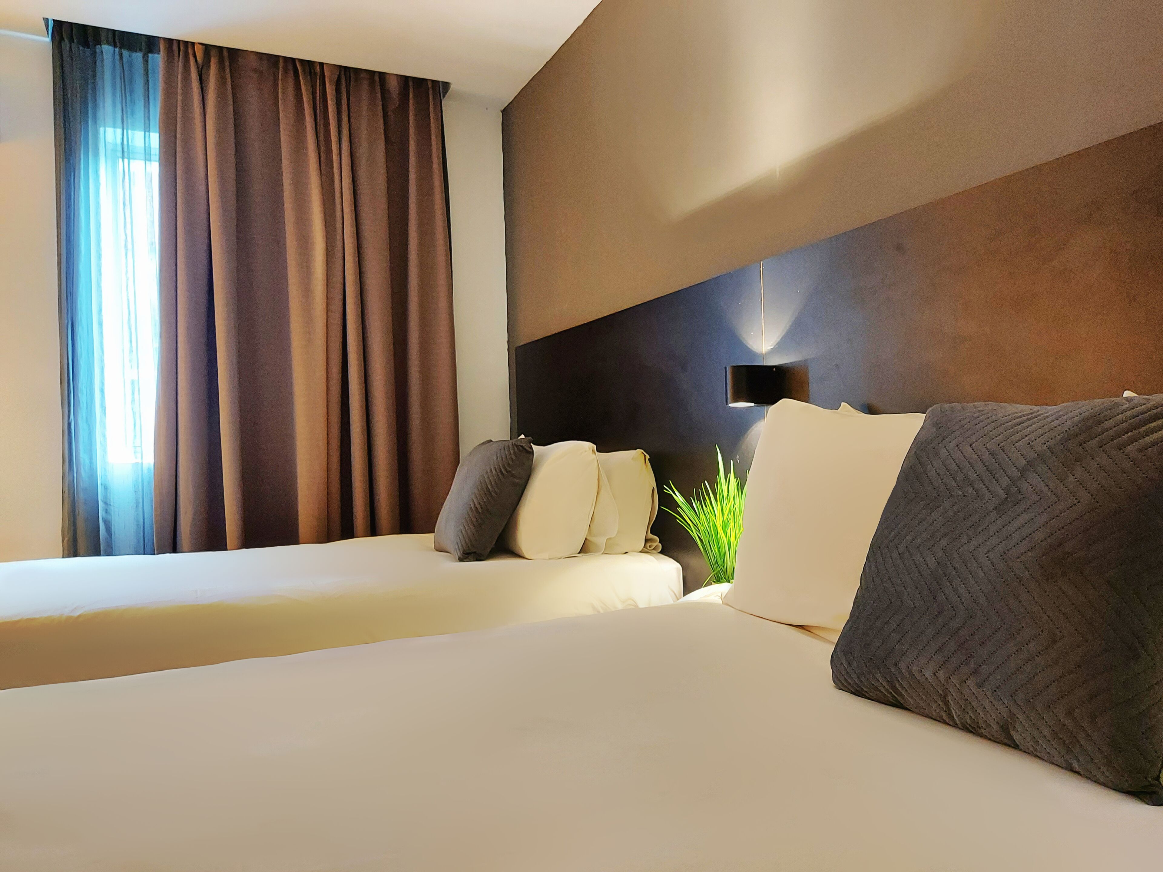 A hotel room with two beds, a large window with curtains, a wall-mounted light, and a plant.