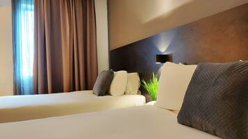 A hotel room with two beds, a large window with curtains, a wall-mounted light, and a plant.