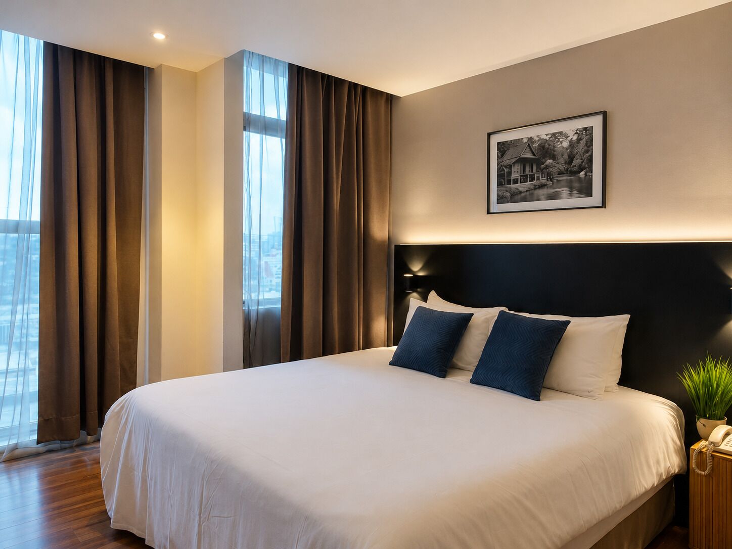 Ein Hotelzimmer mit einem großen Bett, einem Kopfteil mit integrierter Beleuchtung, einem gerahmten Bild an der Wand und Blick auf die Stadt durch das Fenster.