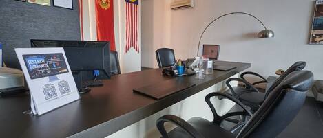 Zona de recepción de hotel con mostrador, computadora, sillas, banderas y certificados en la pared.