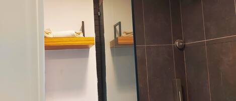 A bathroom with dark tiled walls, a shower with a glass door, and a wooden shelf with towels.
