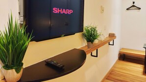 A modern living room with a mounted TV displaying "SHARP" on the screen, a remote control on a black table, a potted plant, and a wooden bench.