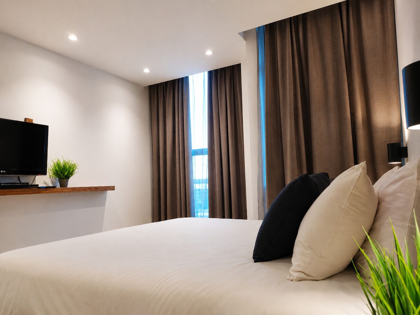 Une chambre d’hôtel moderne avec un grand lit, un téléviseur à écran plat, une petite plante et une vue sur la ville par la fenêtre.